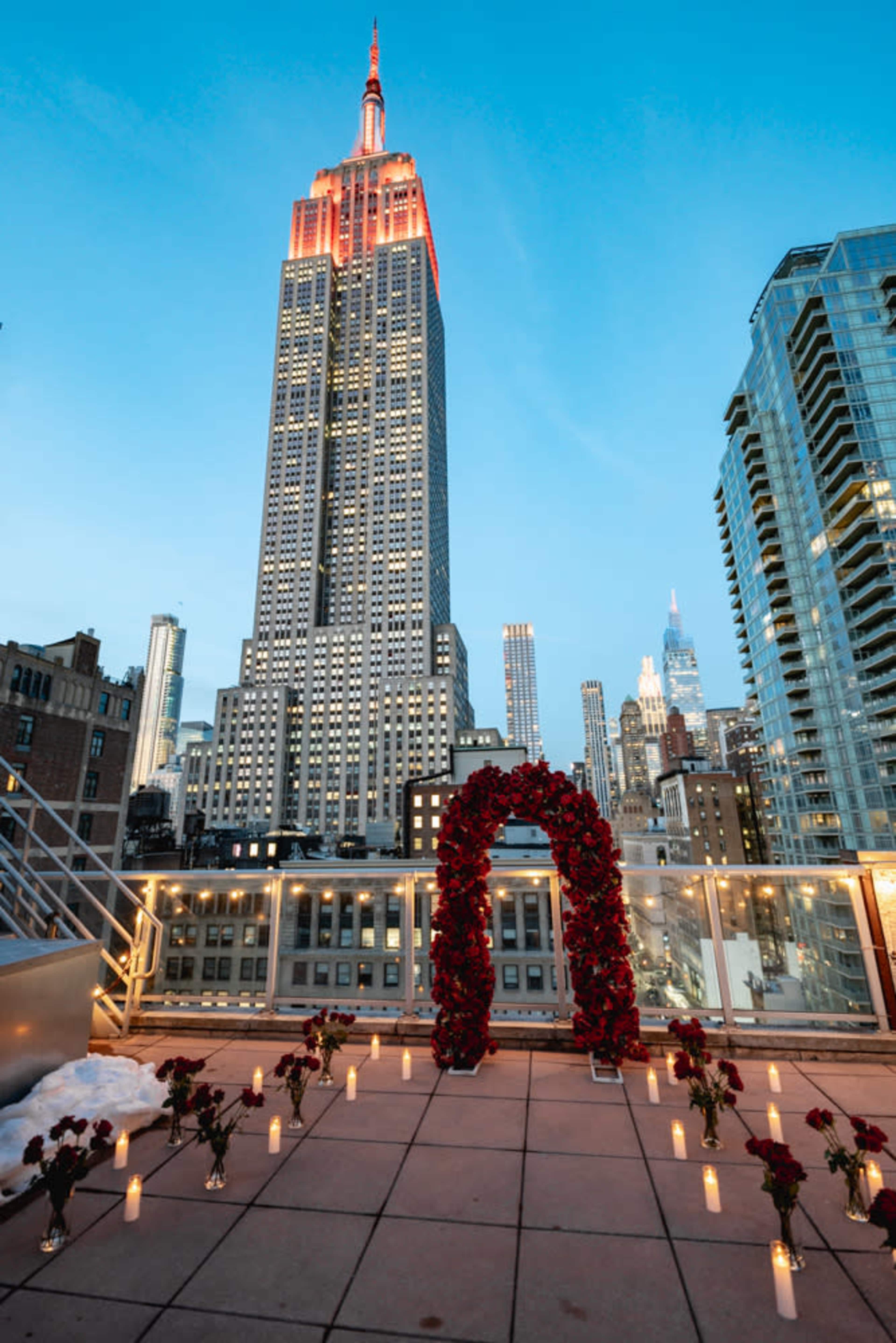 The image shows a rooftop setting adorned with a floral arch and candles, overlooking the Empire State Building illuminated at dusk.