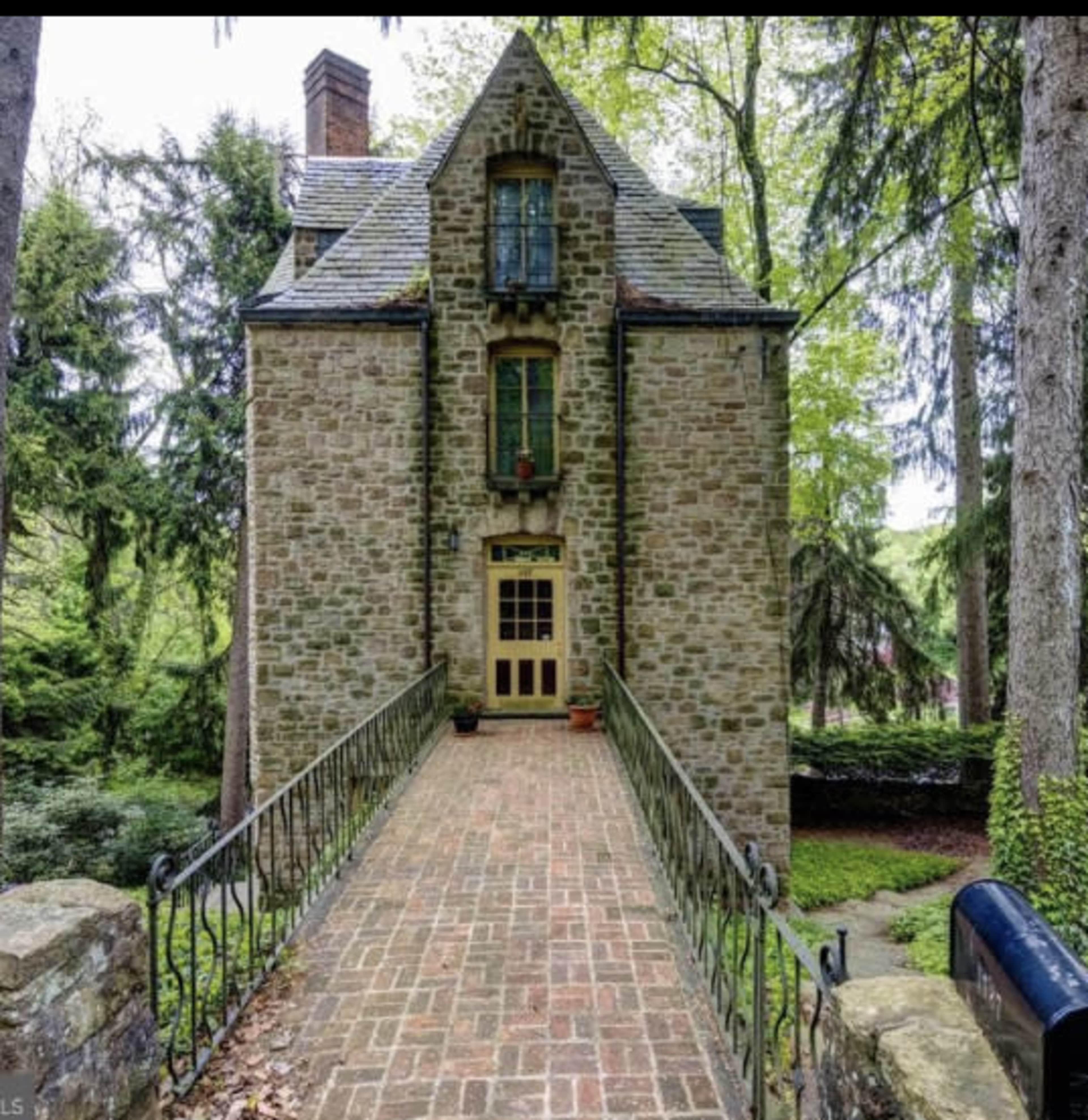 A stone house with a steep roof stands at the end of a brick walkway, surrounded by tall trees.