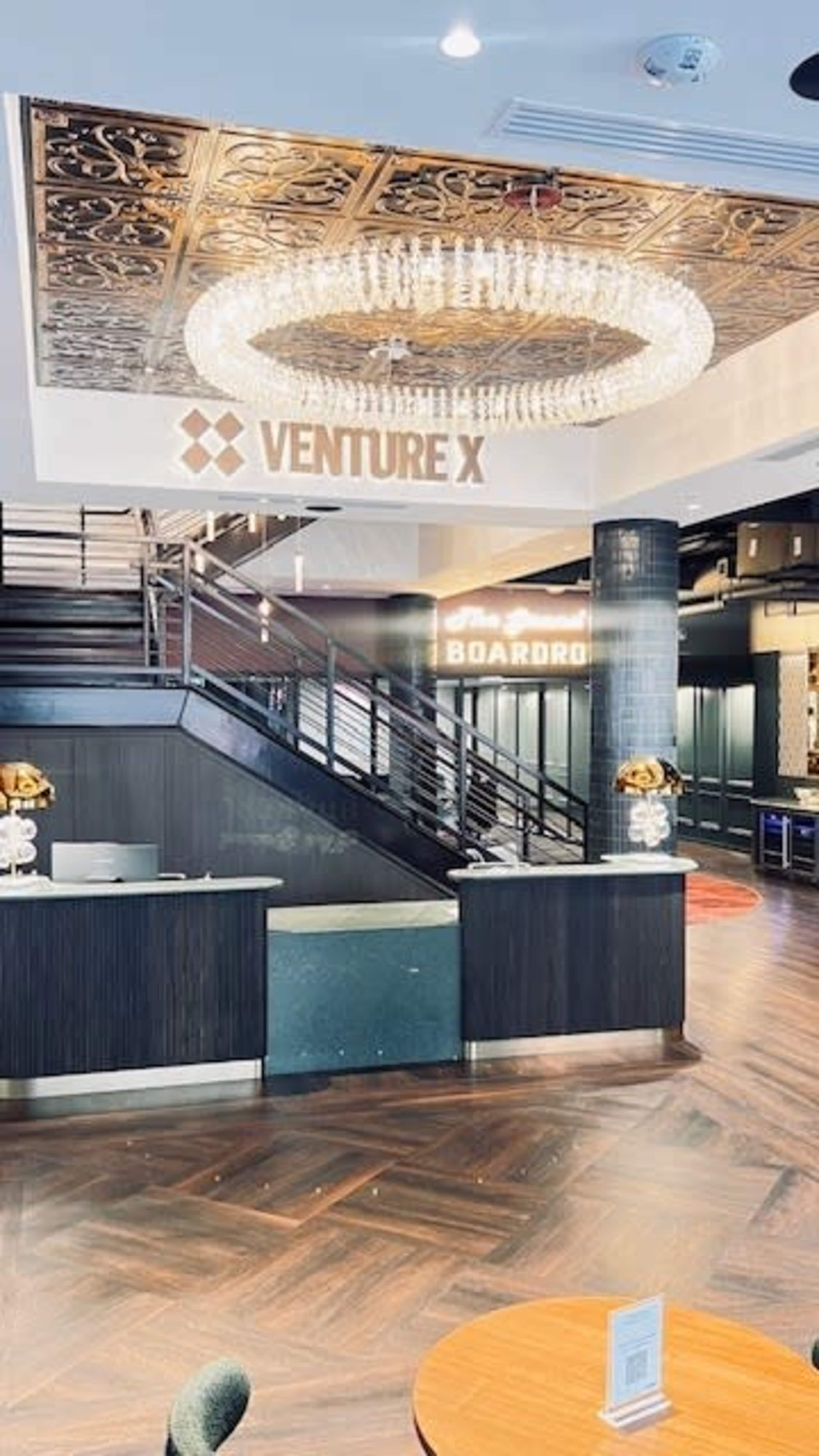 The image shows the interior of a modern business center, featuring a reception area with a sleek desk, a large chandelier, and signs for "Venture X" and "Boardroom" on the walls.