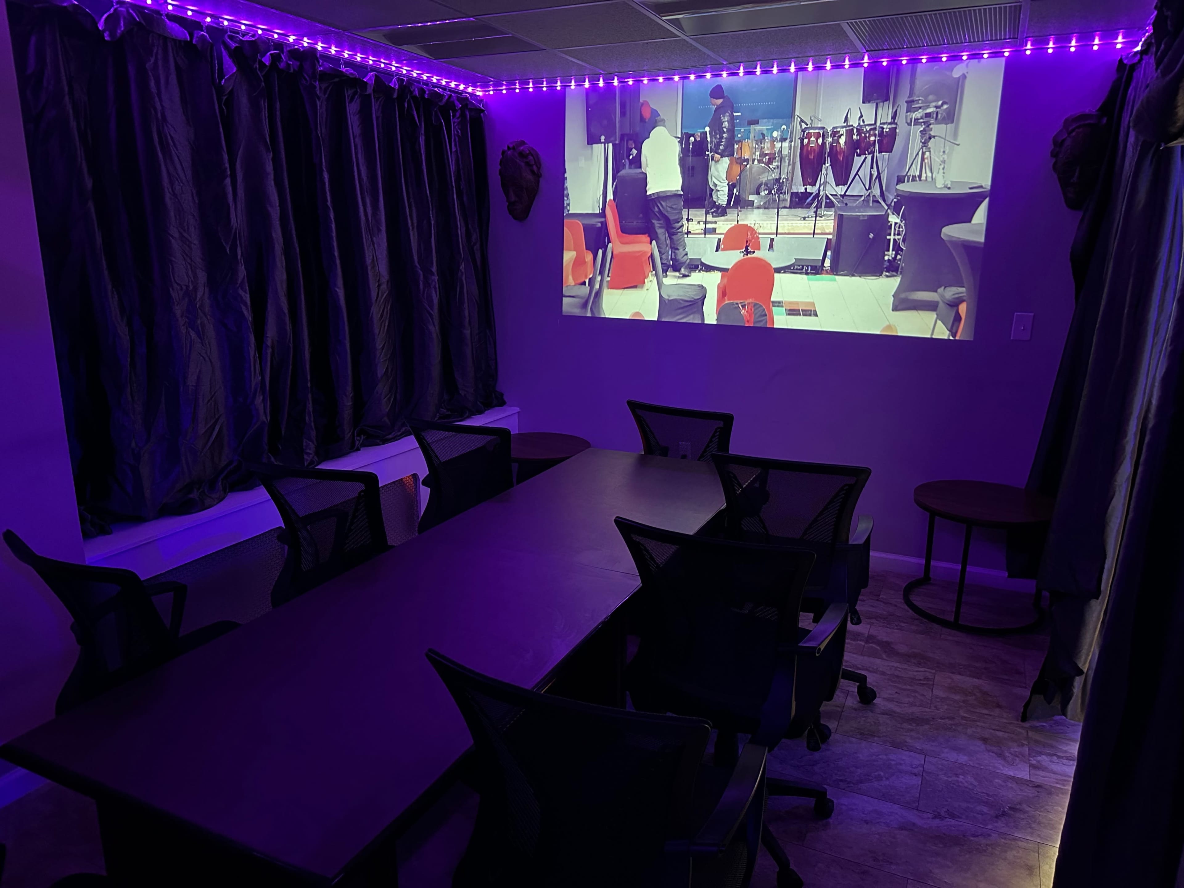 A dimly lit meeting room with a long black table surrounded by black chairs and a projection screen displaying a performance.