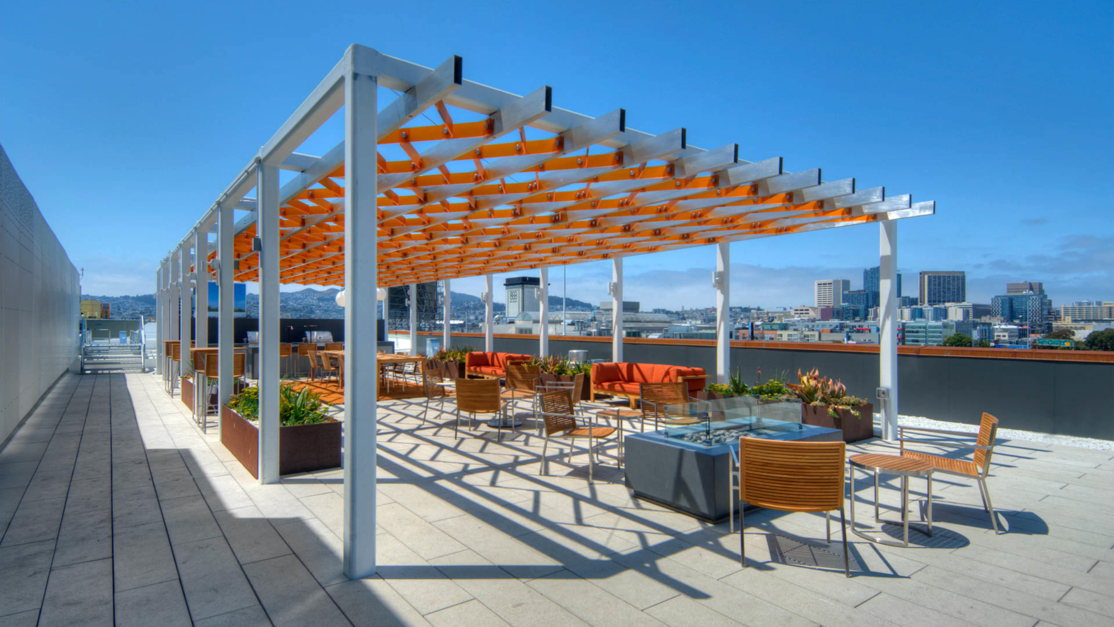 A rooftop terrace features a pergola with orange accents, surrounded by tables and chairs, offering a view of city buildings and a clear sky.