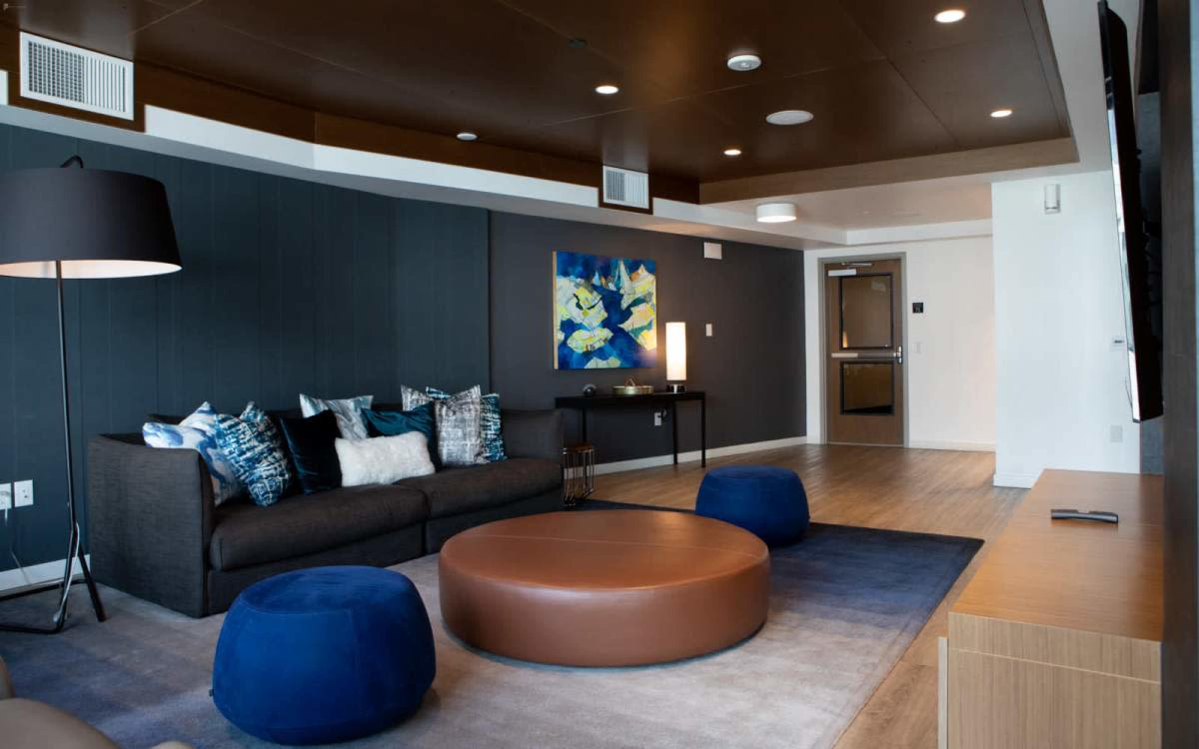 The image shows a modern living room with a dark sofa, round coffee table, and colorful artwork on the wall, complemented by a hardwood floor and ambient lighting.
