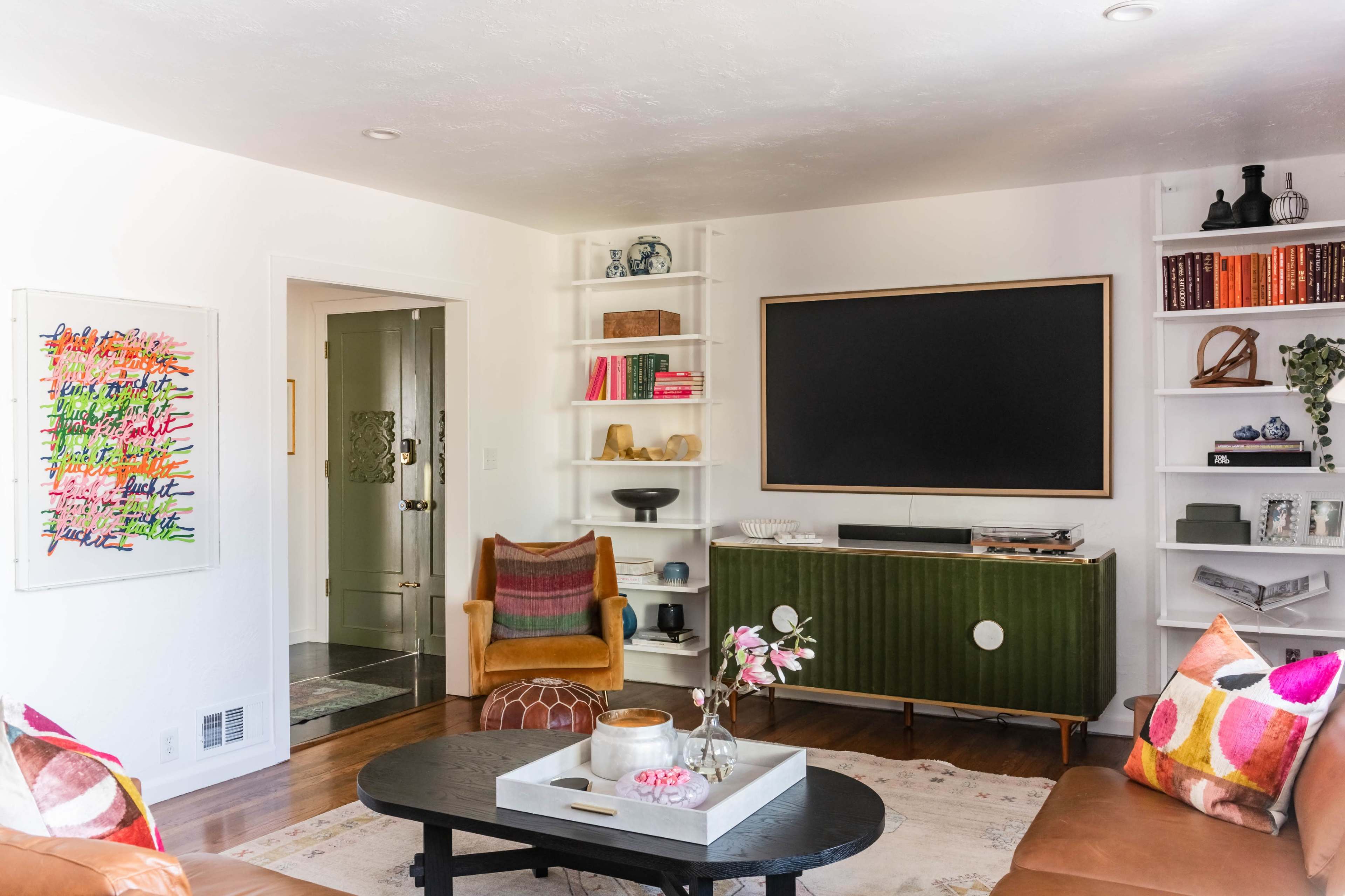 The living room features a black screen mounted on a green entertainment unit, surrounded by bookshelves filled with decor and books, and a central coffee table adorned with decorative items.
