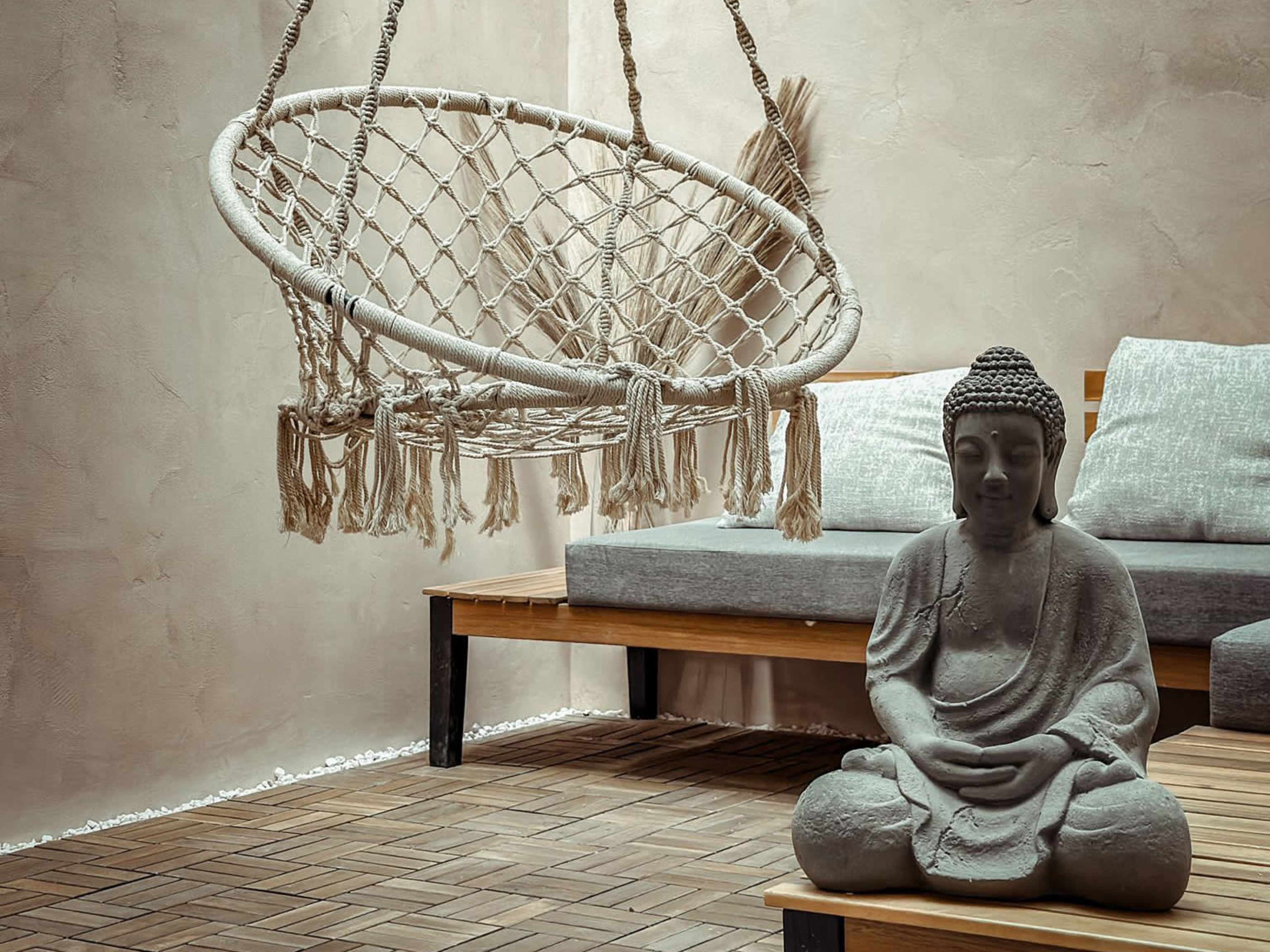 A hanging macramé chair is positioned next to a seated Buddha statue on a wooden floor in a minimalist living space.