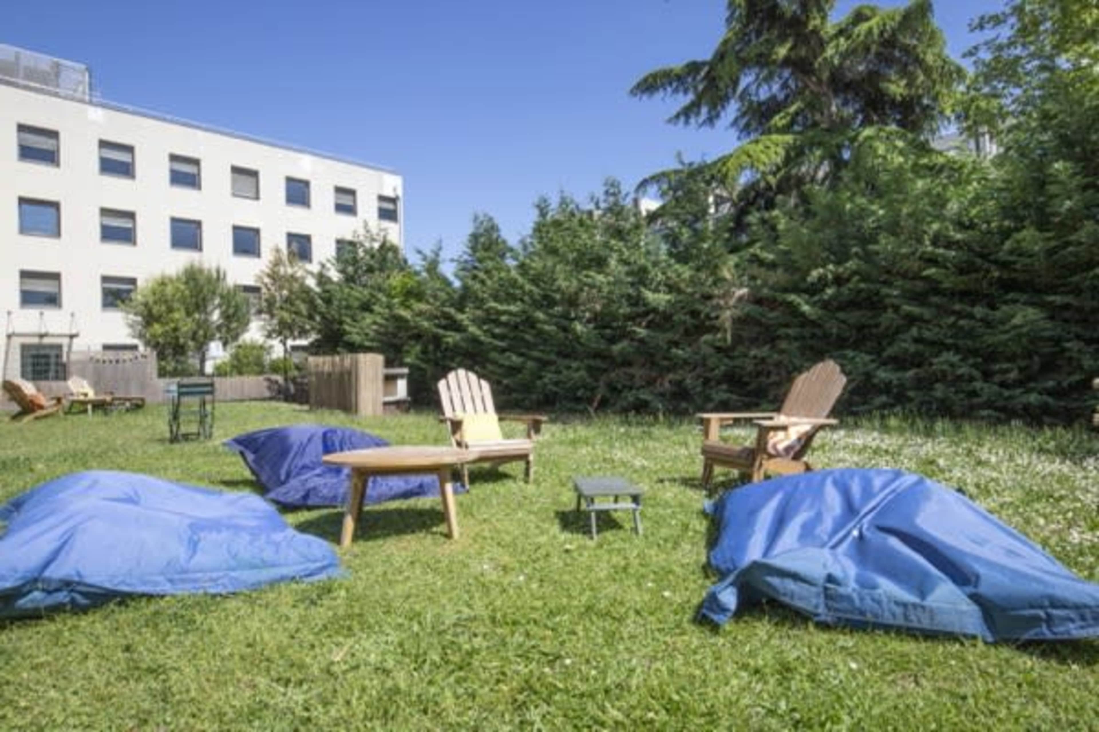 A garden area with several blue beanbags, a table, and two wooden chairs surrounded by bushes and a building in the background.