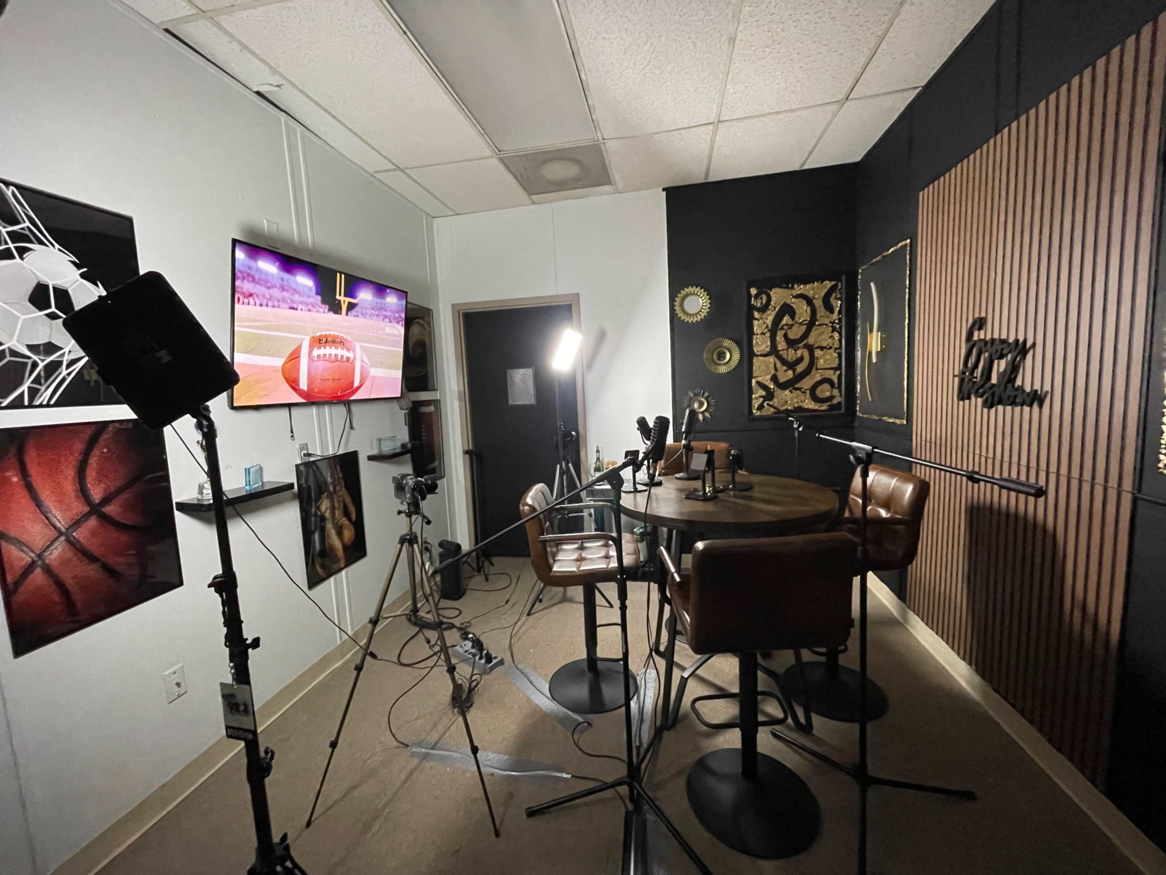 The image shows a small studio setup with a circular table and four chairs, surrounded by various sports-themed wall art and equipped with cameras and lighting for recording.