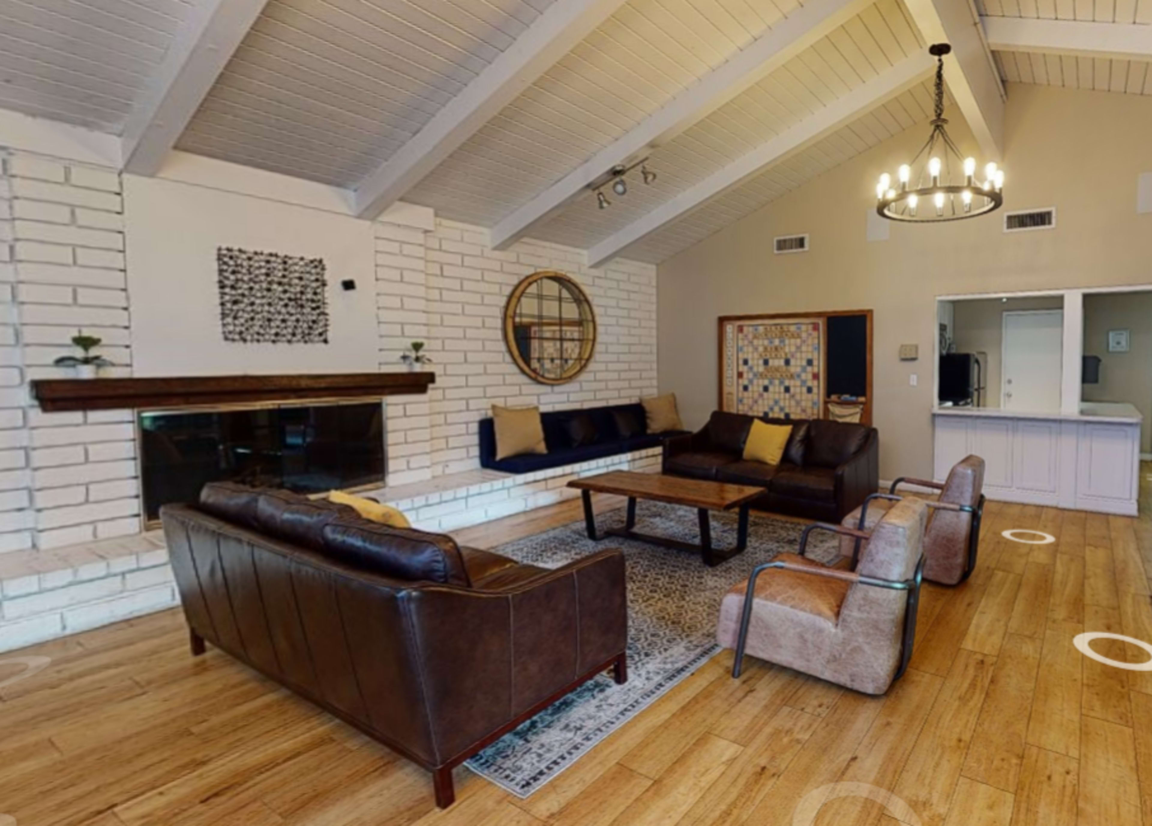 A modern living room with a brown leather sofa, two armchairs, a wooden coffee table, and a fireplace, set against a backdrop of white brick walls and wooden ceiling beams.