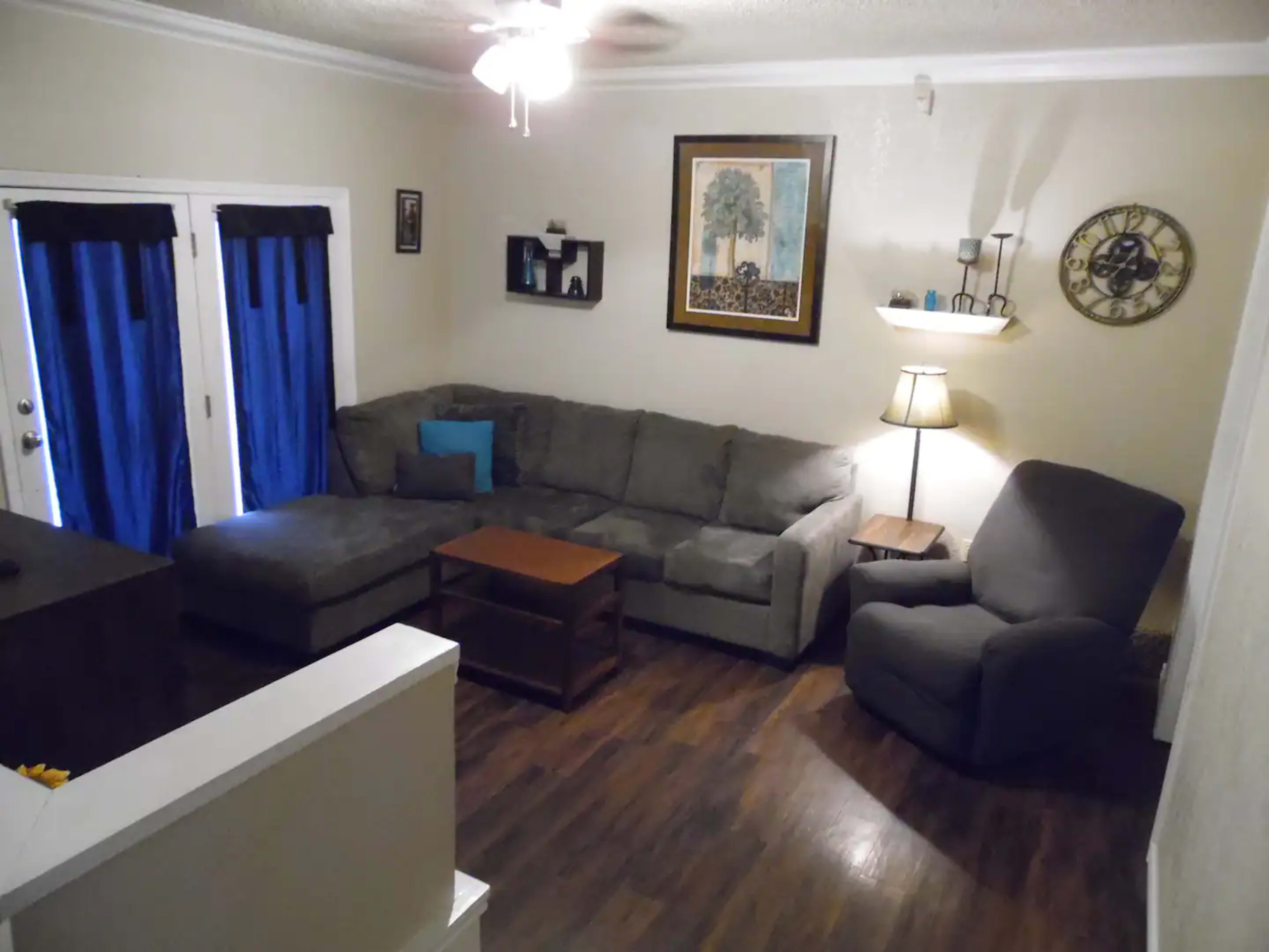 A living room featuring a sectional sofa, a wooden coffee table, a lamp, and a wall clock, with blue curtains covering the windows.