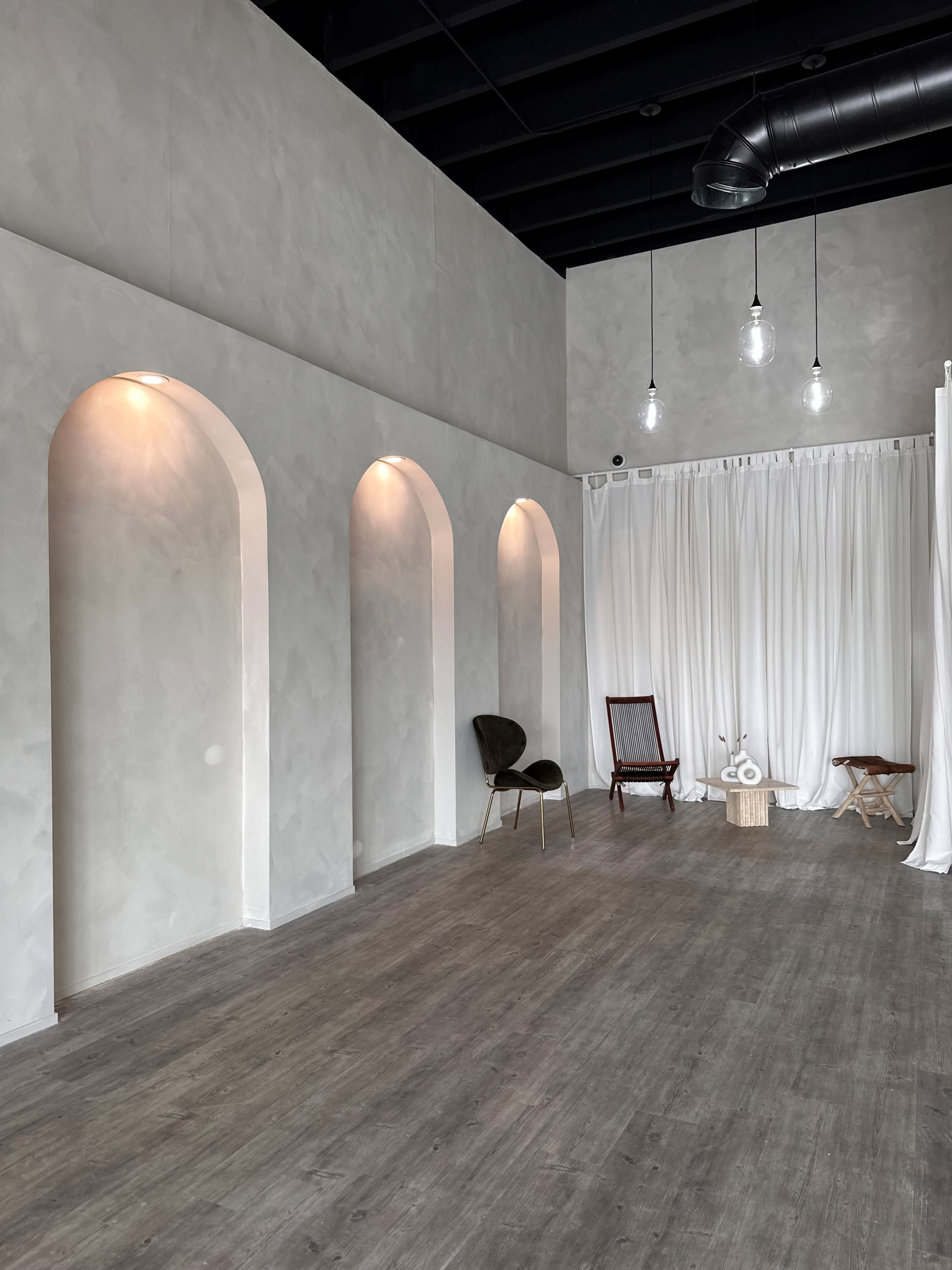 The image shows a spacious, minimalist room with arched wall niches, a white curtain, and a mix of wooden and upholstered furniture.