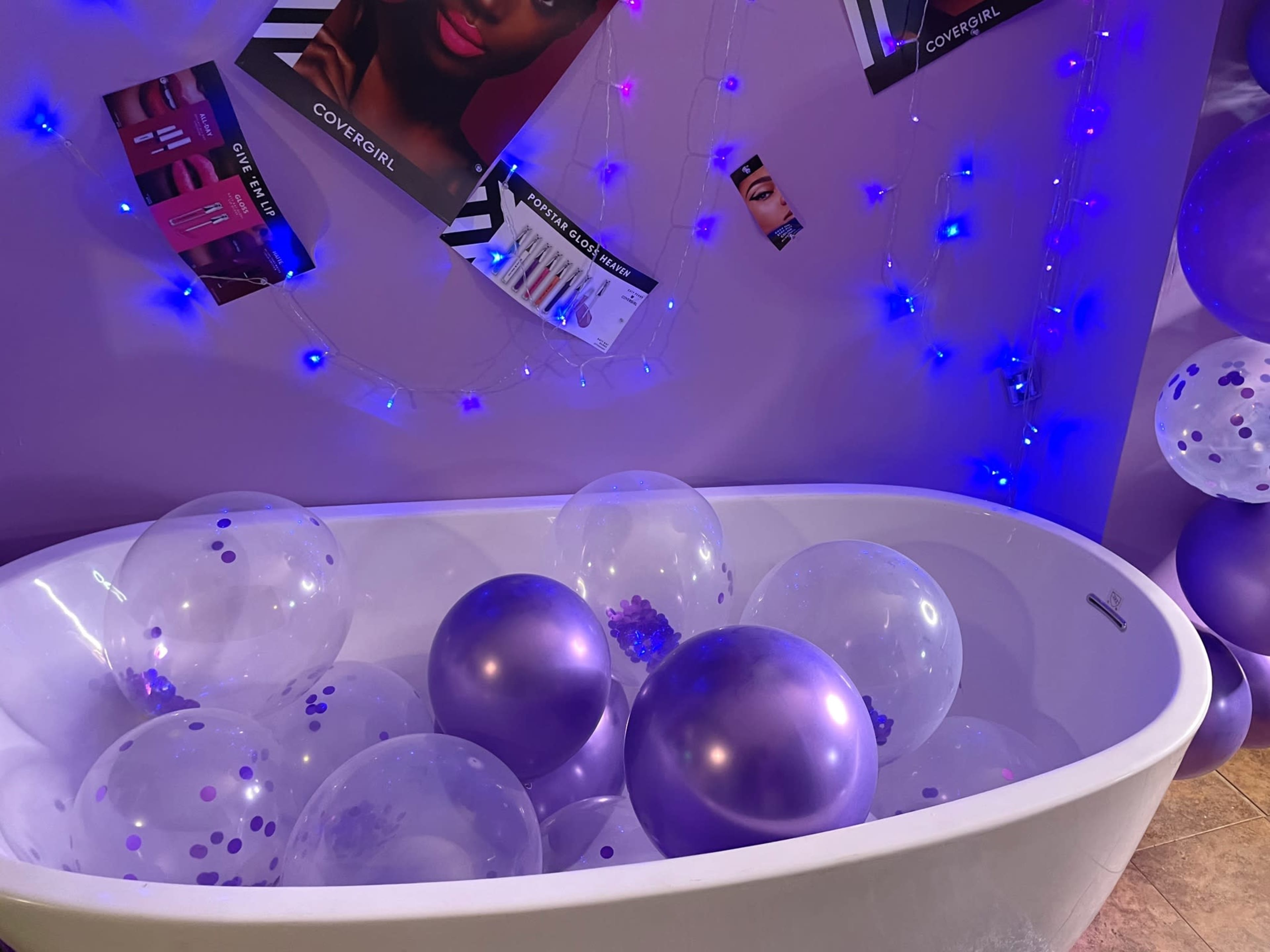 A white bathtub filled with clear and purple balloons is positioned against a lavender wall adorned with promotional posters and string lights.