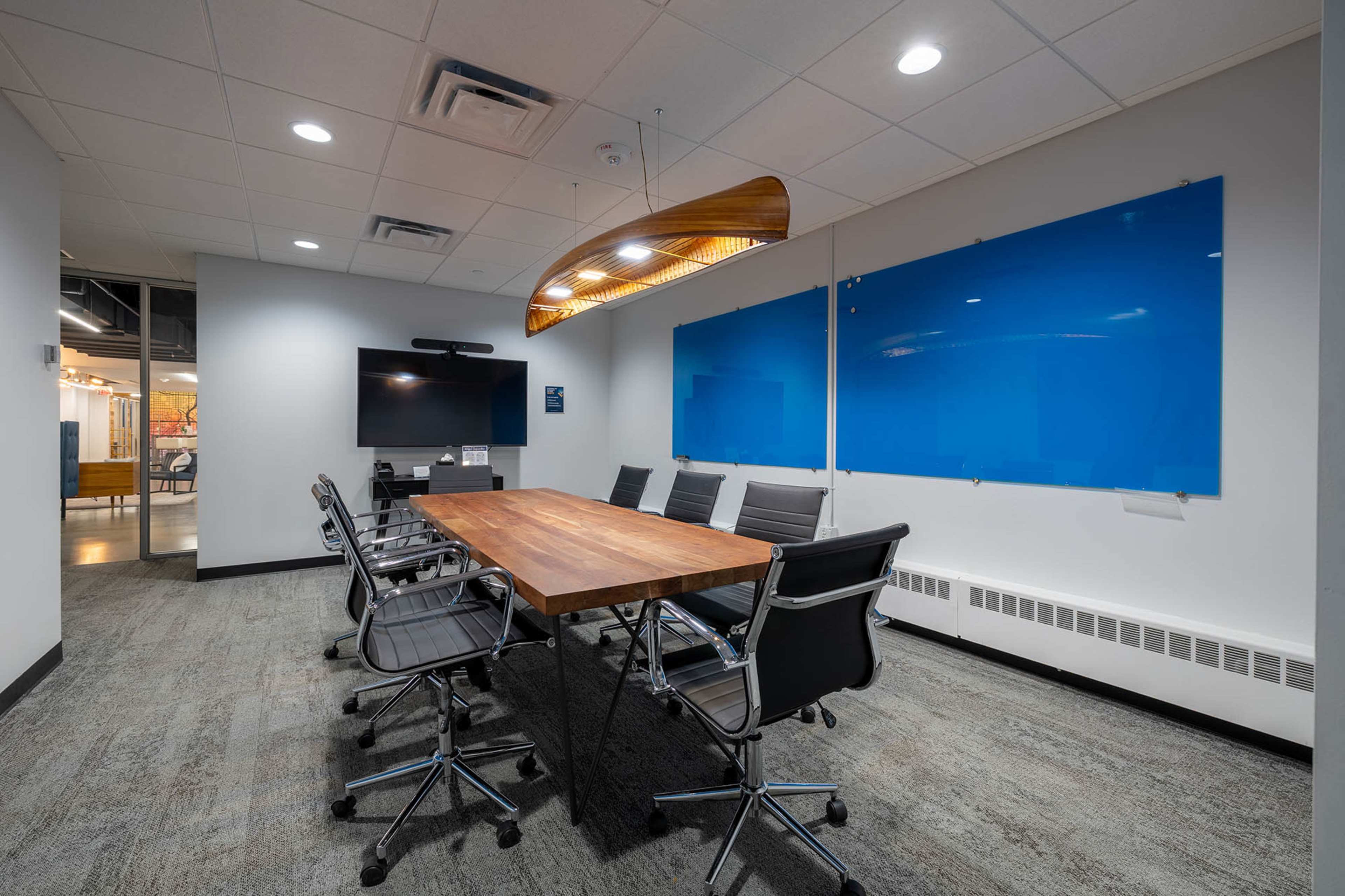 The image shows a modern conference room with a large wooden table surrounded by black ergonomic chairs and blue wall panels.