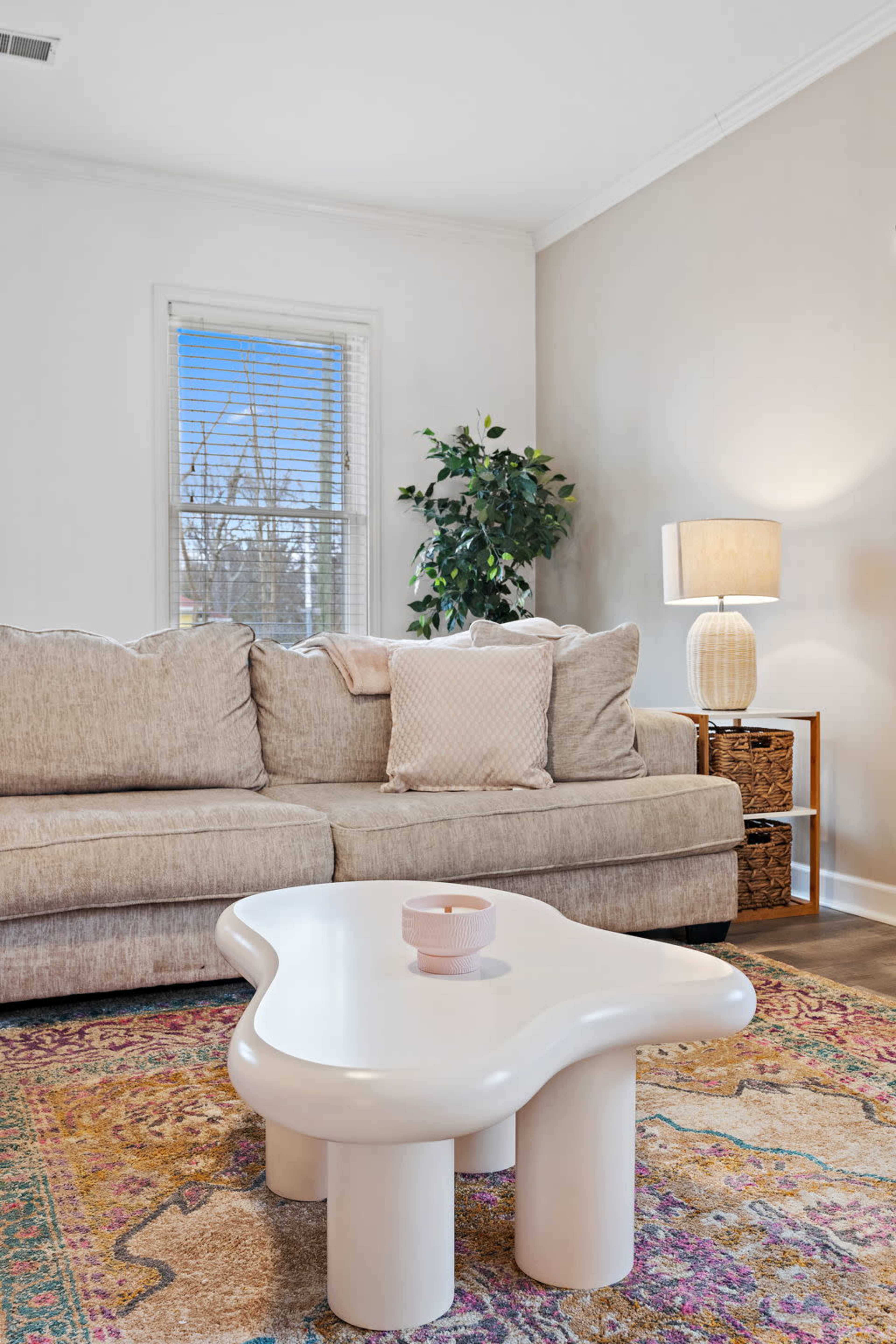 A light-colored sofa is positioned in a living room with a patterned rug, a round table, a floor lamp, and a decorative plant in the corner.