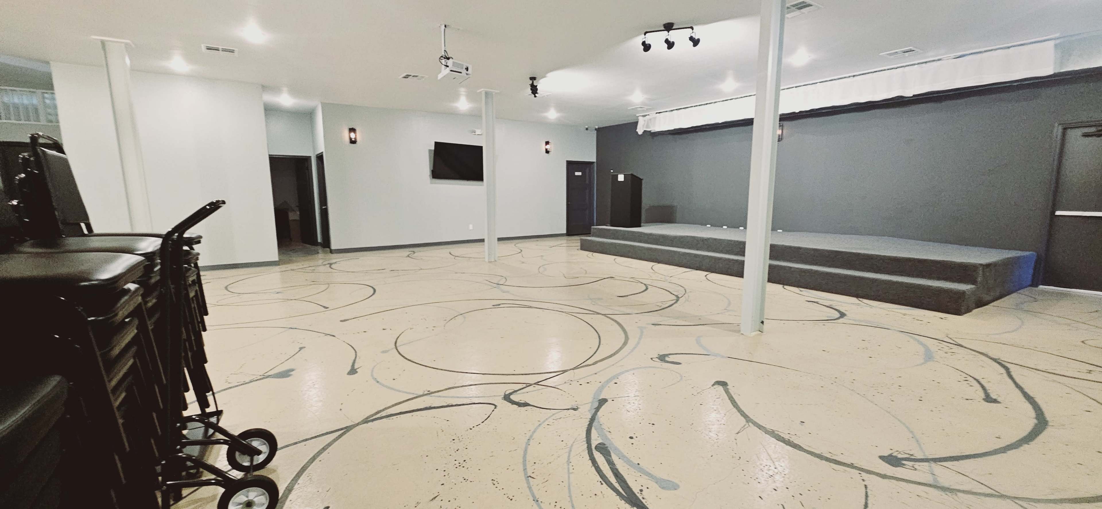 A spacious indoor venue with a stage at one end, gray walls, a polished floor featuring swirling patterns, and stacked chairs against the wall.