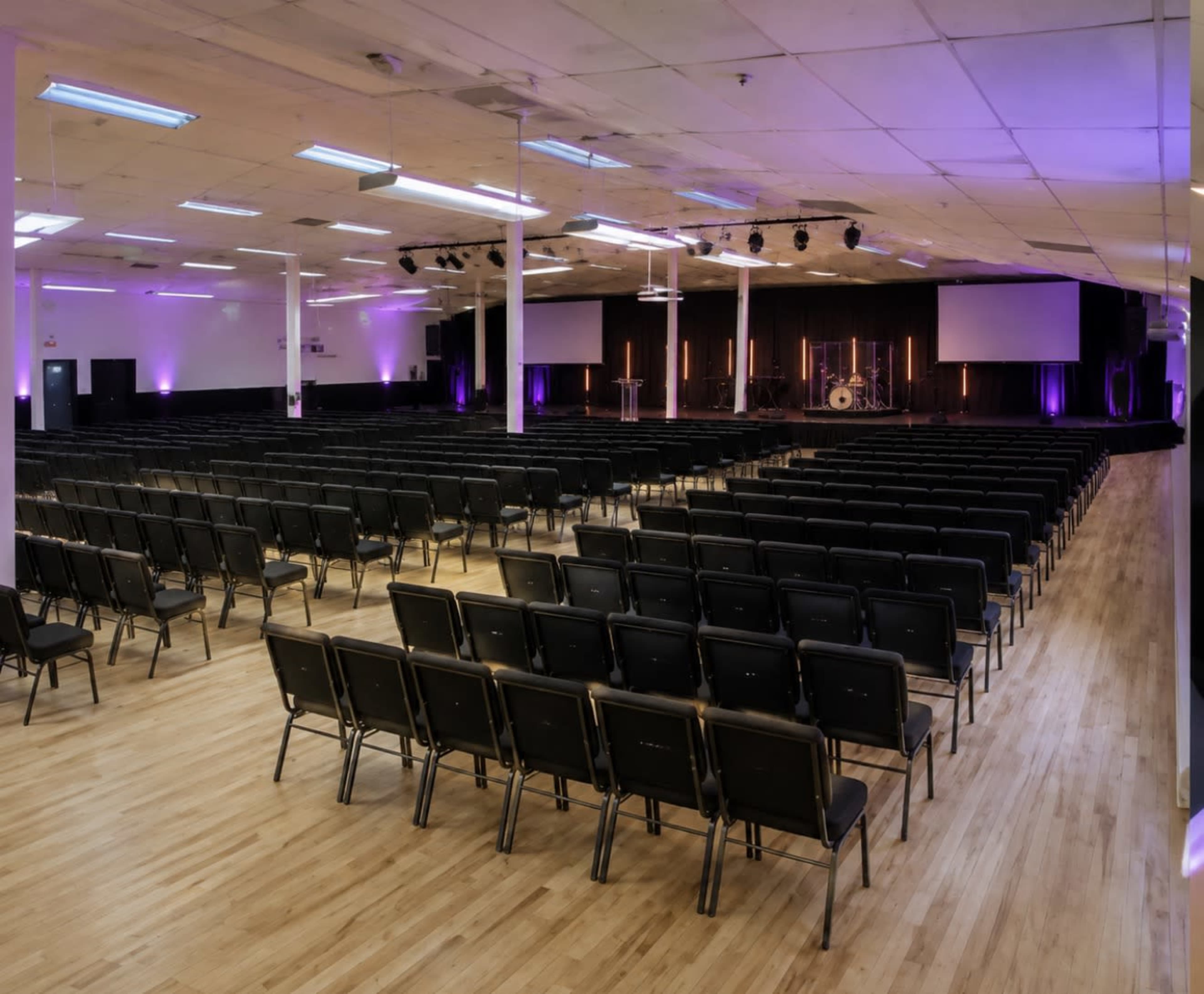 The image shows a large auditorium filled with rows of black folding chairs, with soft purple lighting and a stage at the front.