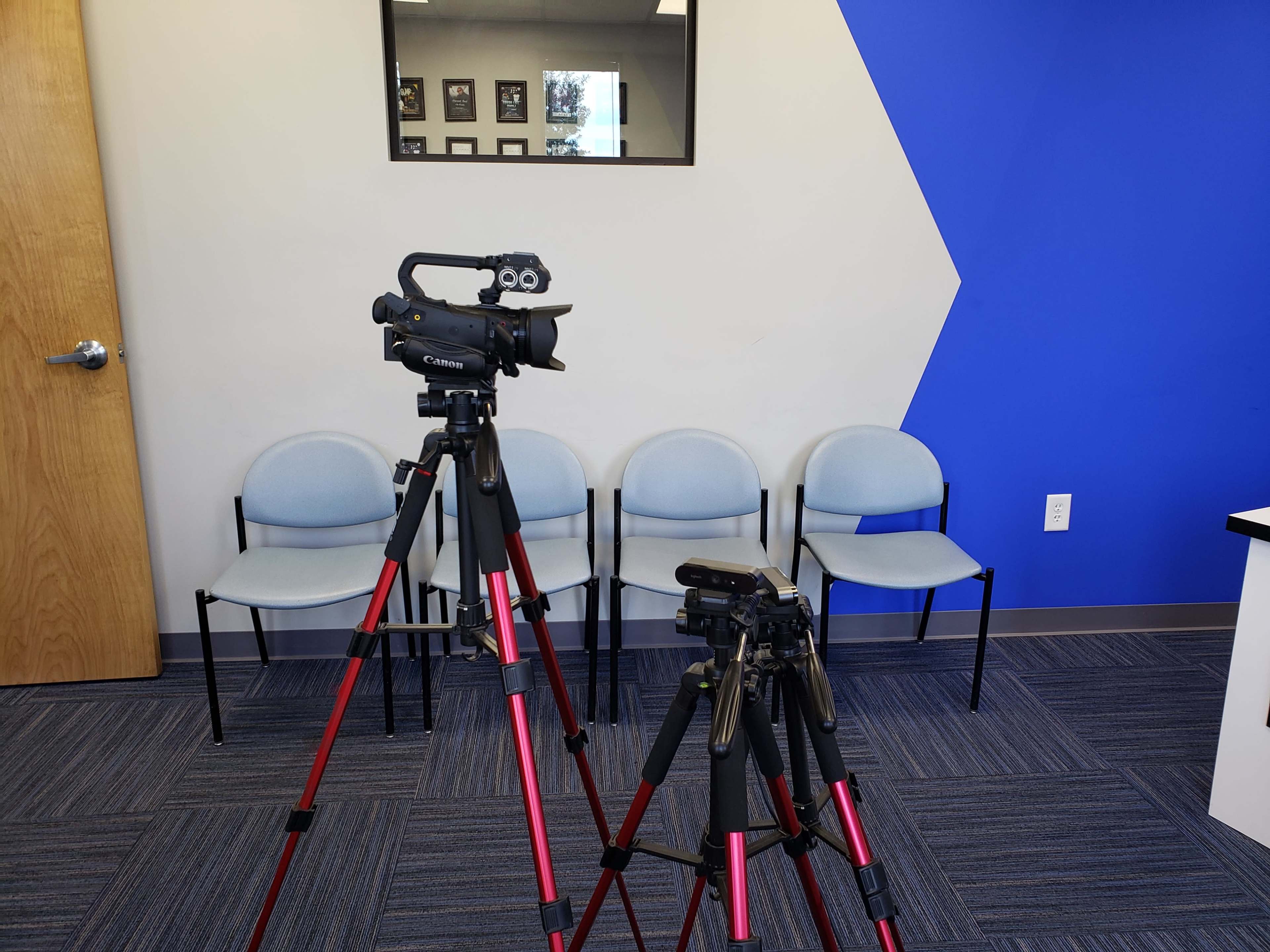 Two tripods with cameras are positioned in an empty room featuring blue and gray walls, with several chairs visible in the background.