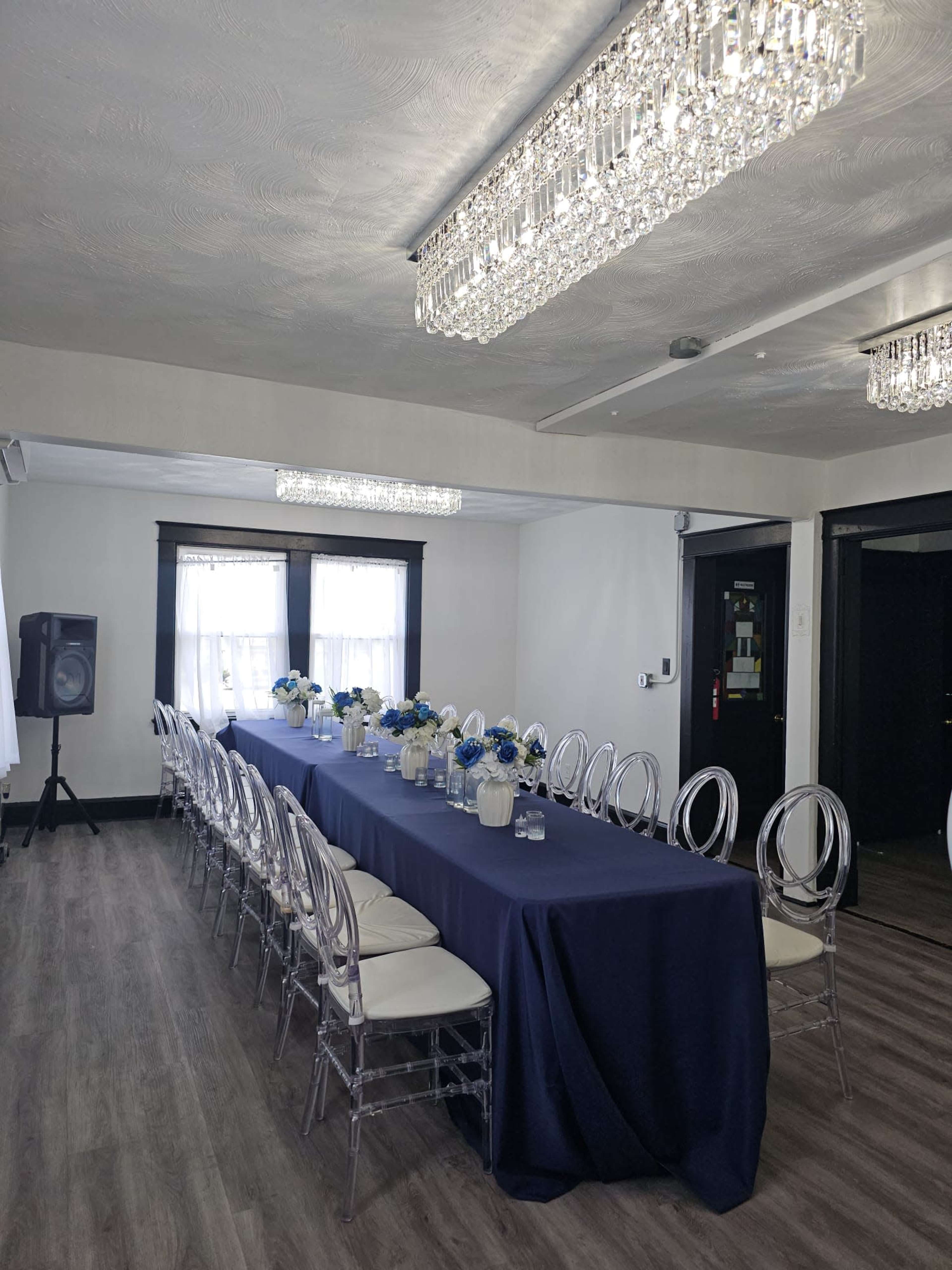 A long, rectangular dining table with a navy blue tablecloth is set in a well-lit room, surrounded by clear acrylic chairs and decorated with floral centerpieces.