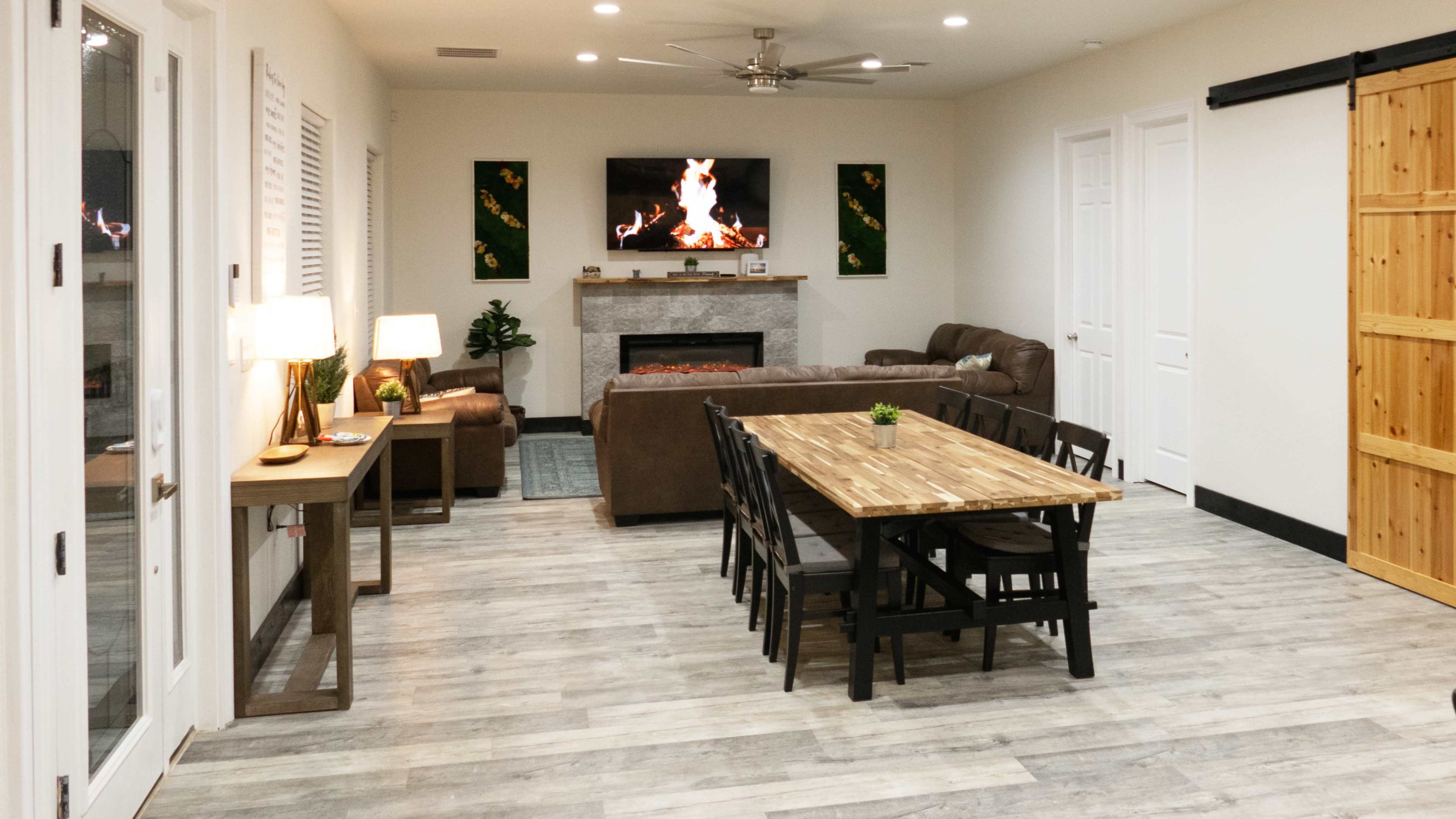A spacious living area featuring a fireplace with a television above it, a brown sofa, a wooden dining table with black chairs, and natural light coming through the windows.