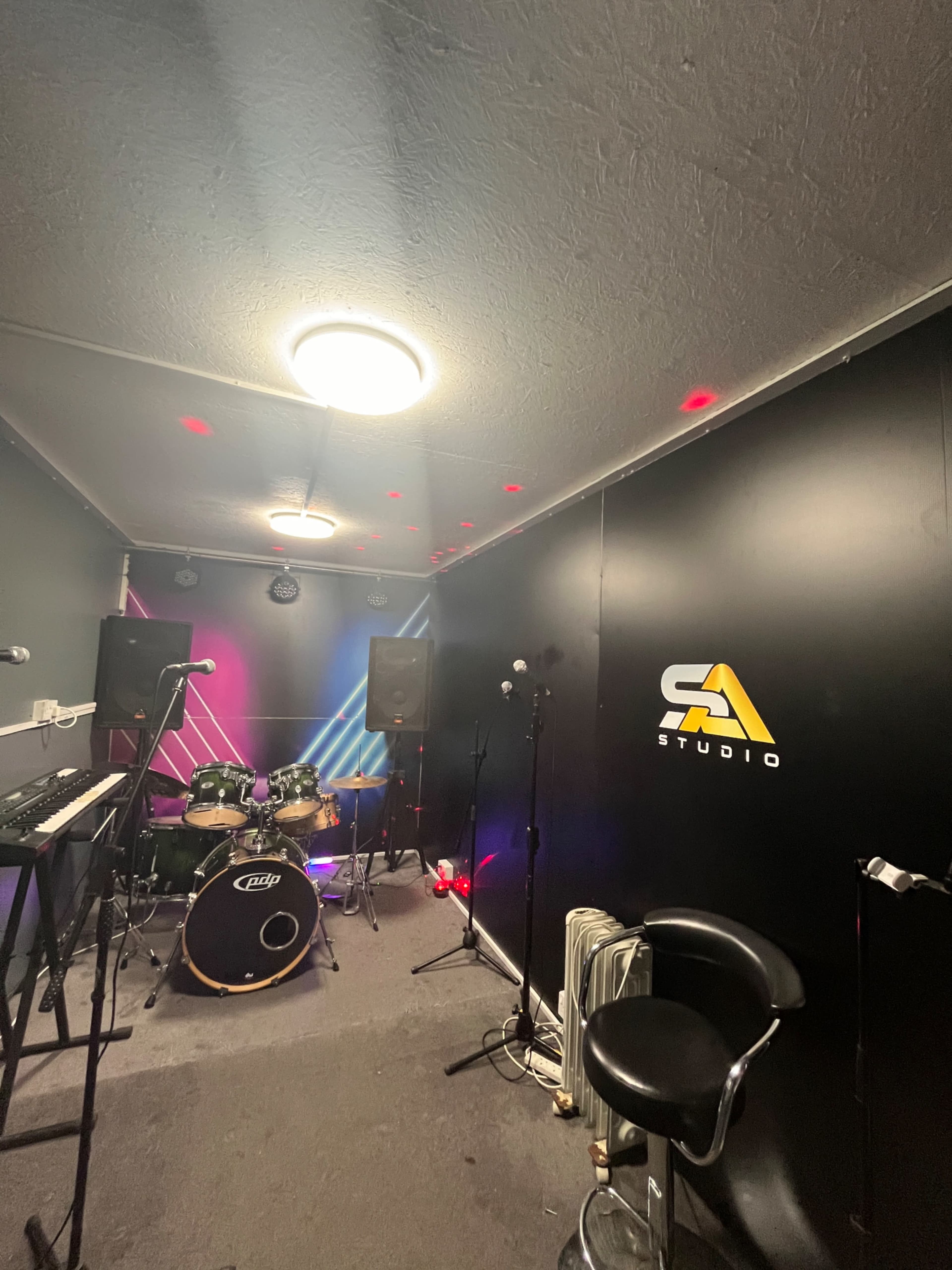 The image shows a small music studio equipped with a drum set, keyboard, and sound equipment, featuring a black wall with a logo and colorful lighting.