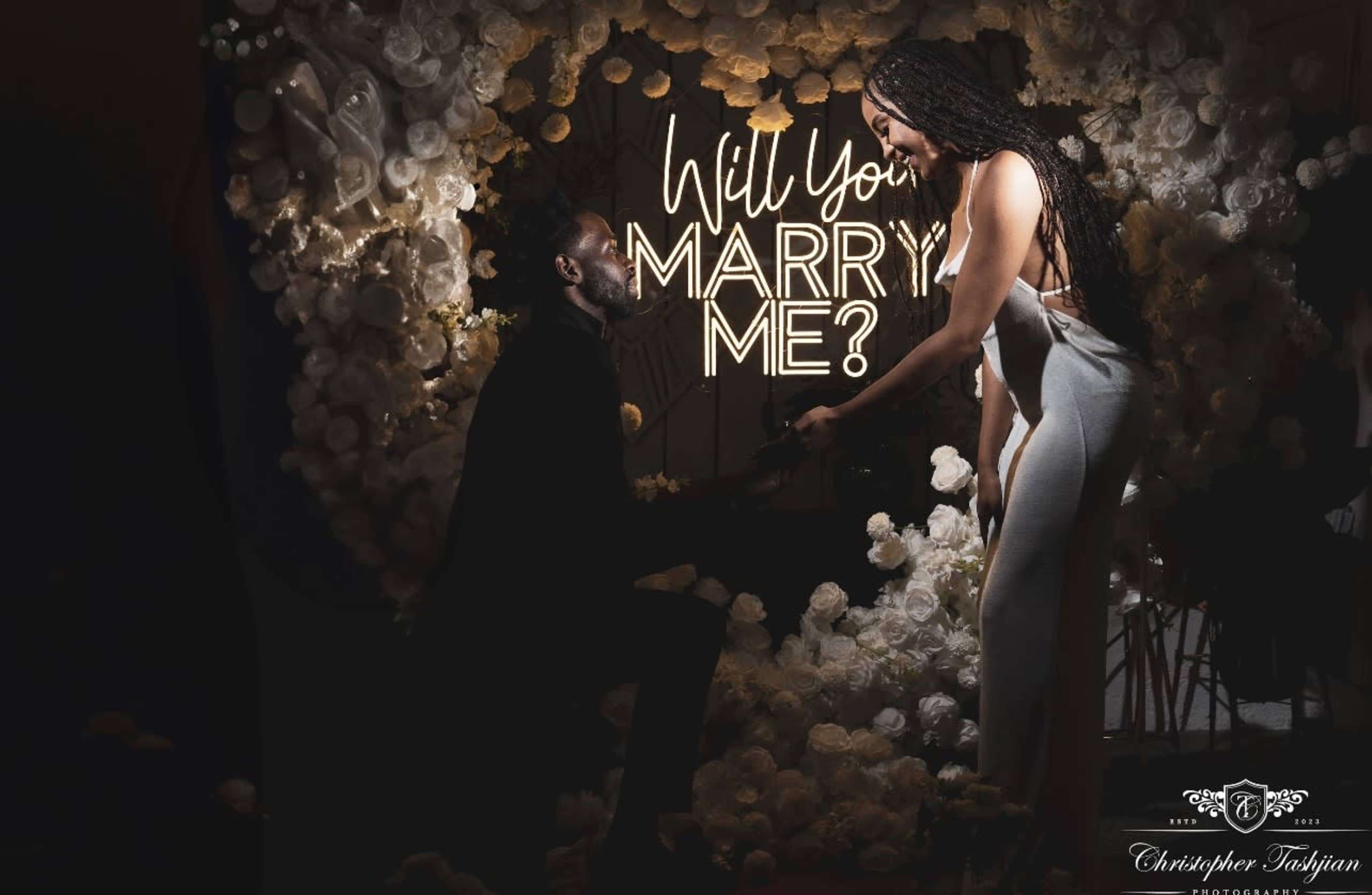 A man is kneeling and presenting a ring to a woman against a background of flowers and the illuminated words "Will You Marry Me?"