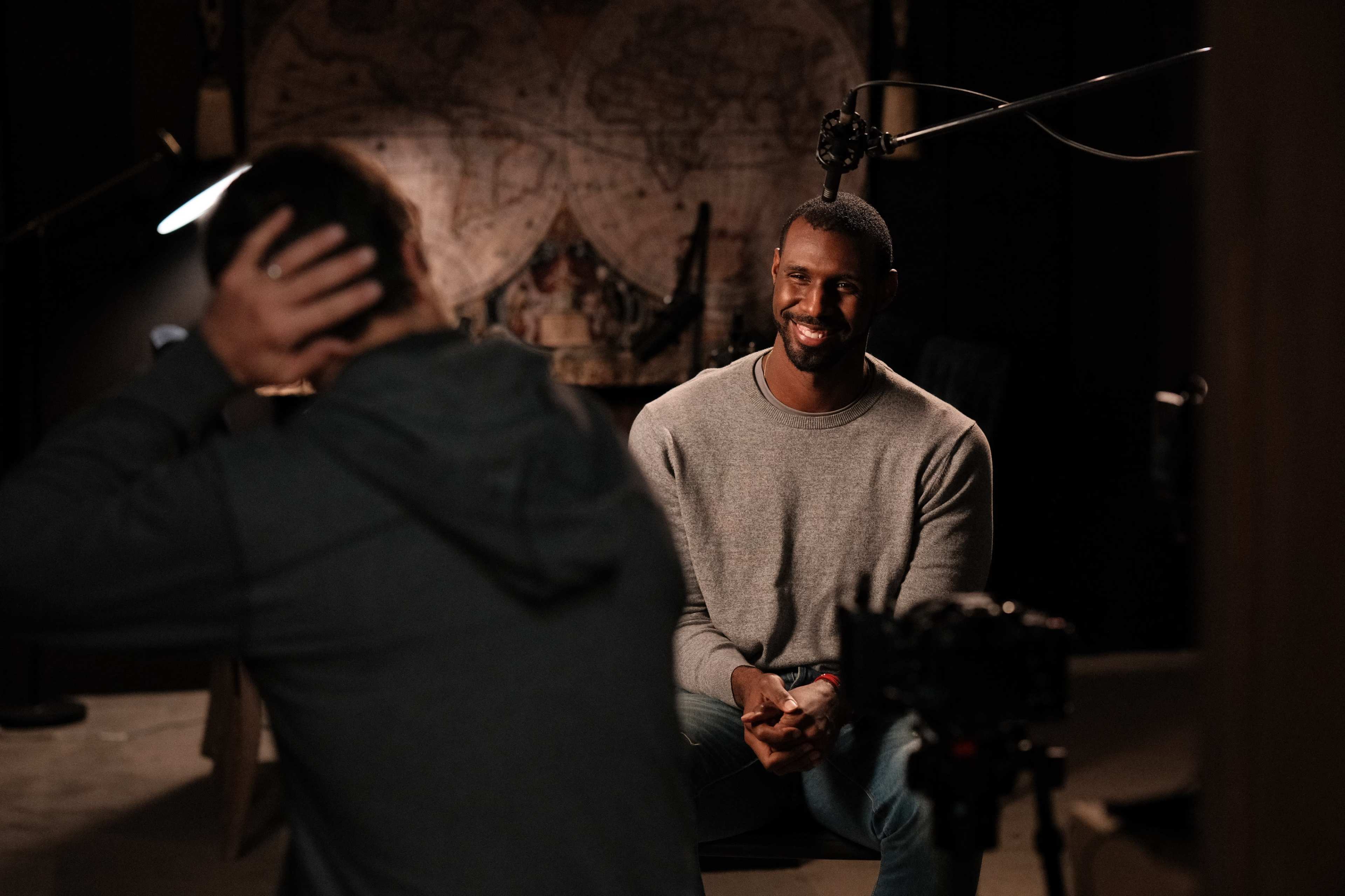 A man in a gray sweater smiles while sitting in front of a camera in a dimly lit room with a map backdrop.