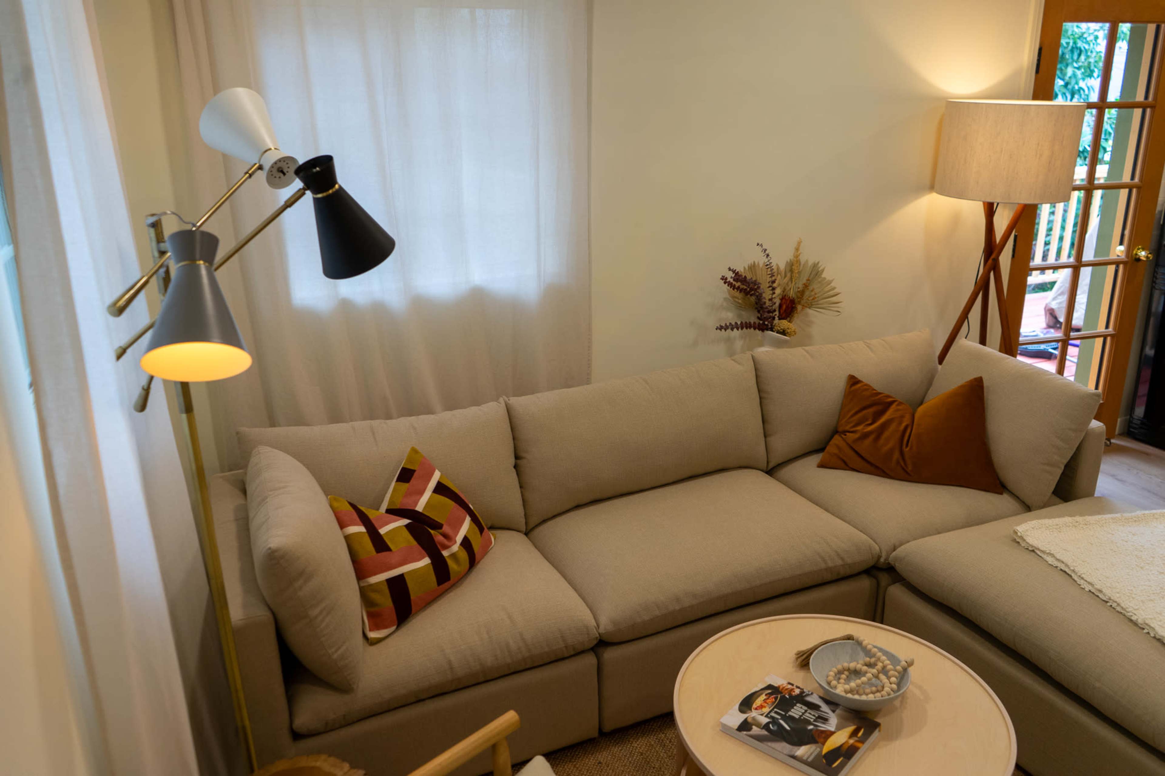 A light-colored, modern living room features a sectional sofa with decorative cushions, a side table with a magazine, and a floor lamp beside a window.