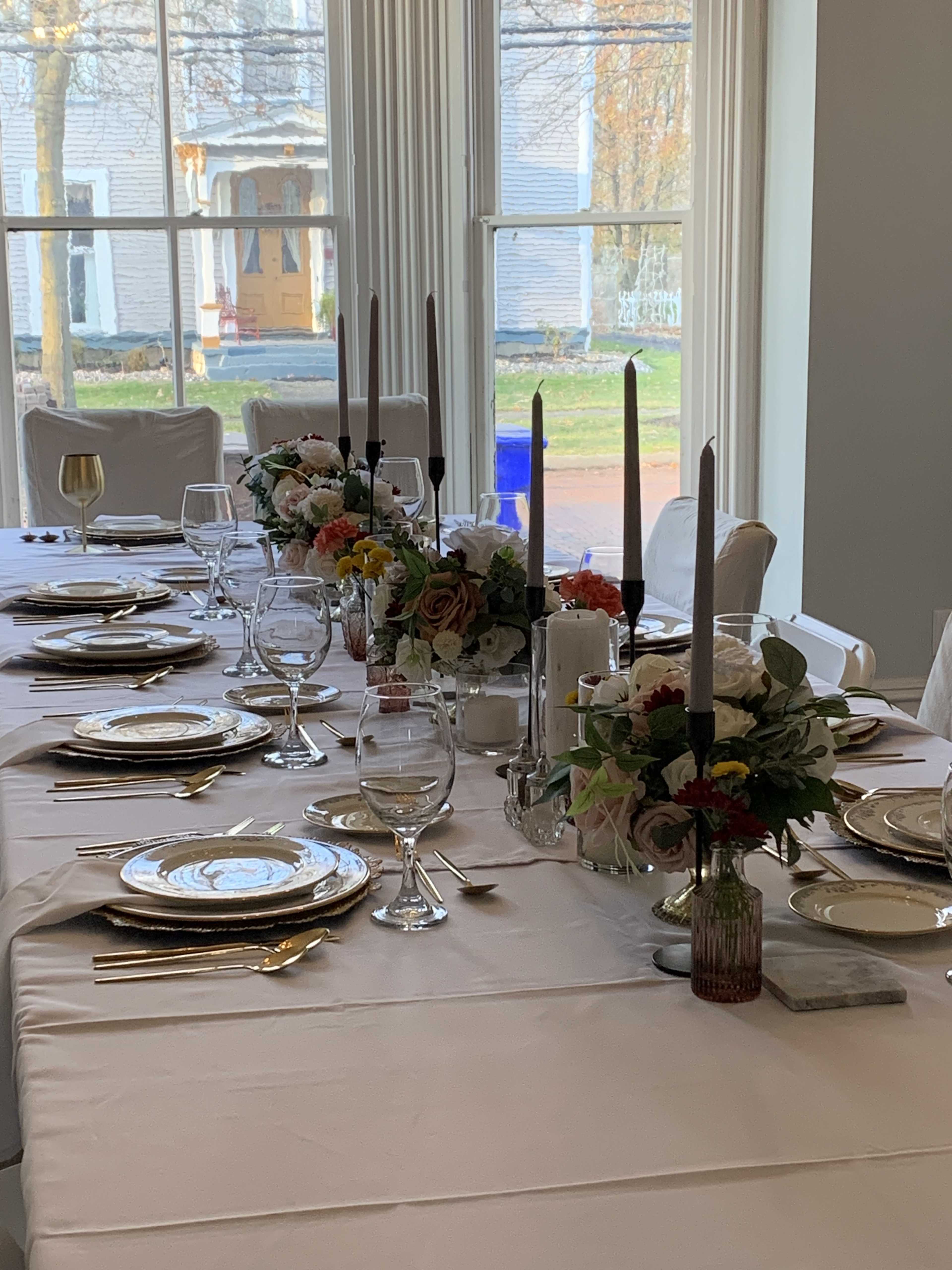 A dining table is set with elegant tableware, including plates, glasses, and candleholders, surrounded by floral arrangements and a soft tablecloth.
