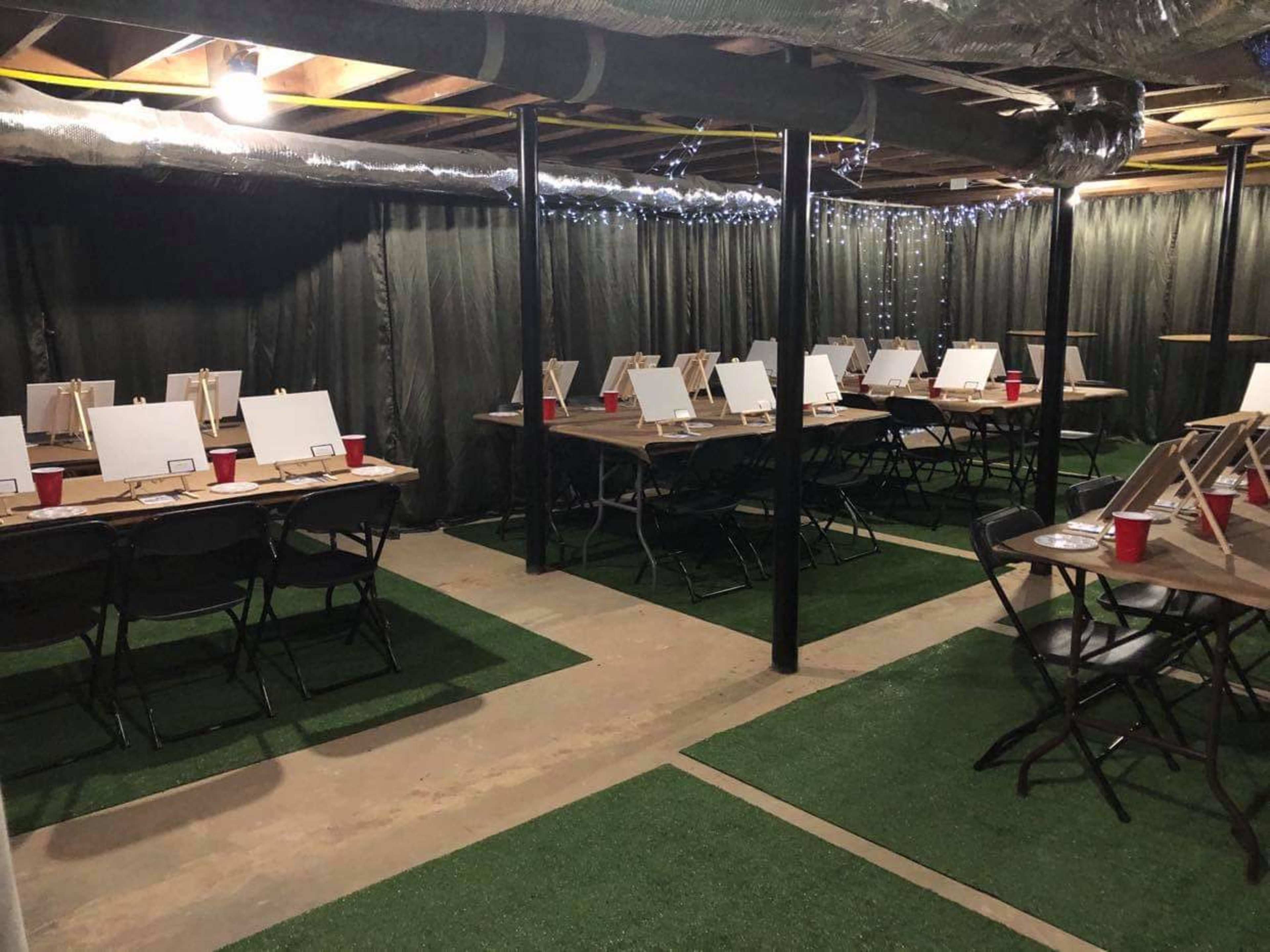 A basement room is set up with tables and chairs, each equipped with blank canvases and red cups.