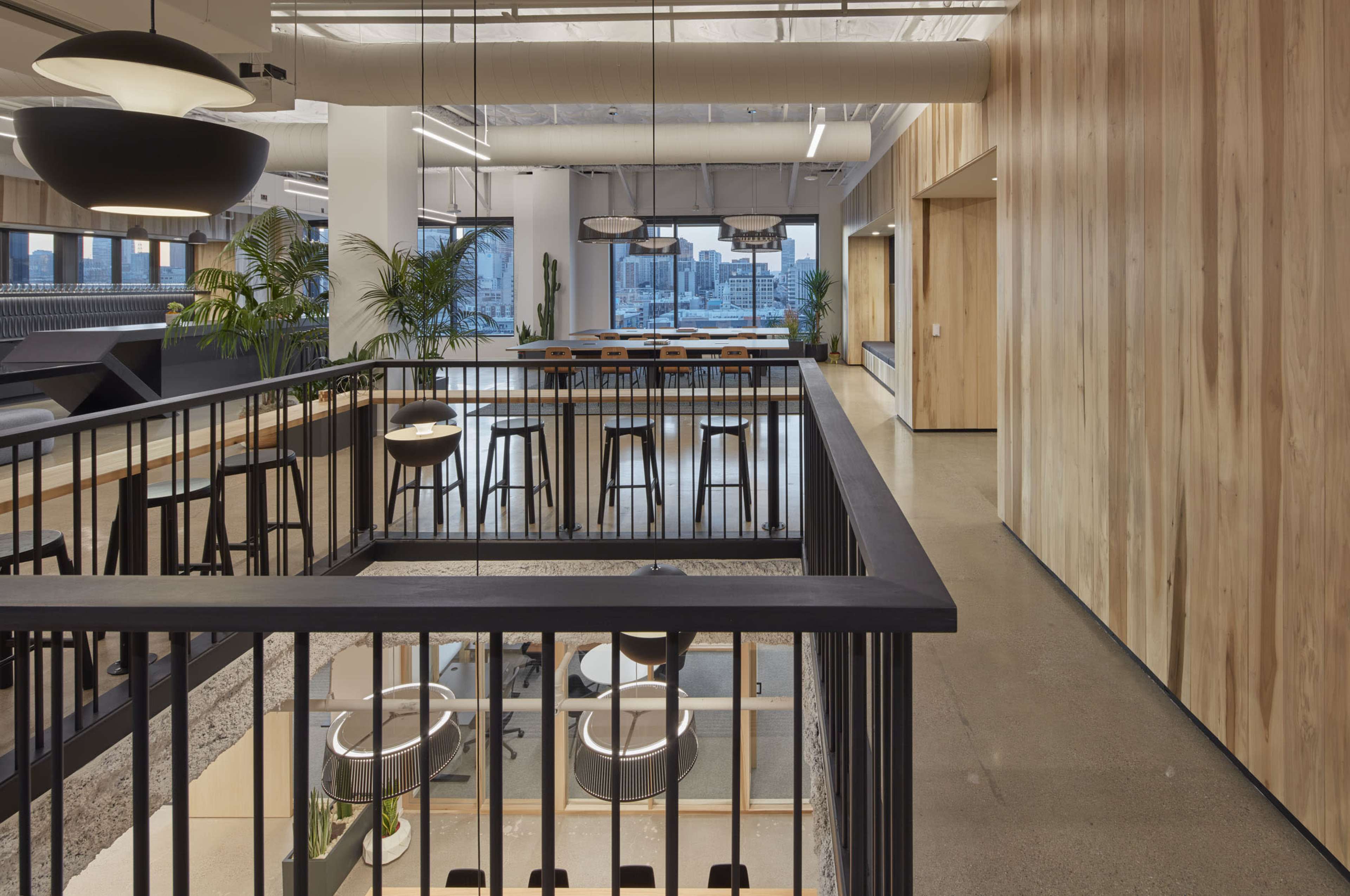 A modern office interior with wooden paneling, a balcony overlooking a workspace, and large windows showcasing a city skyline.