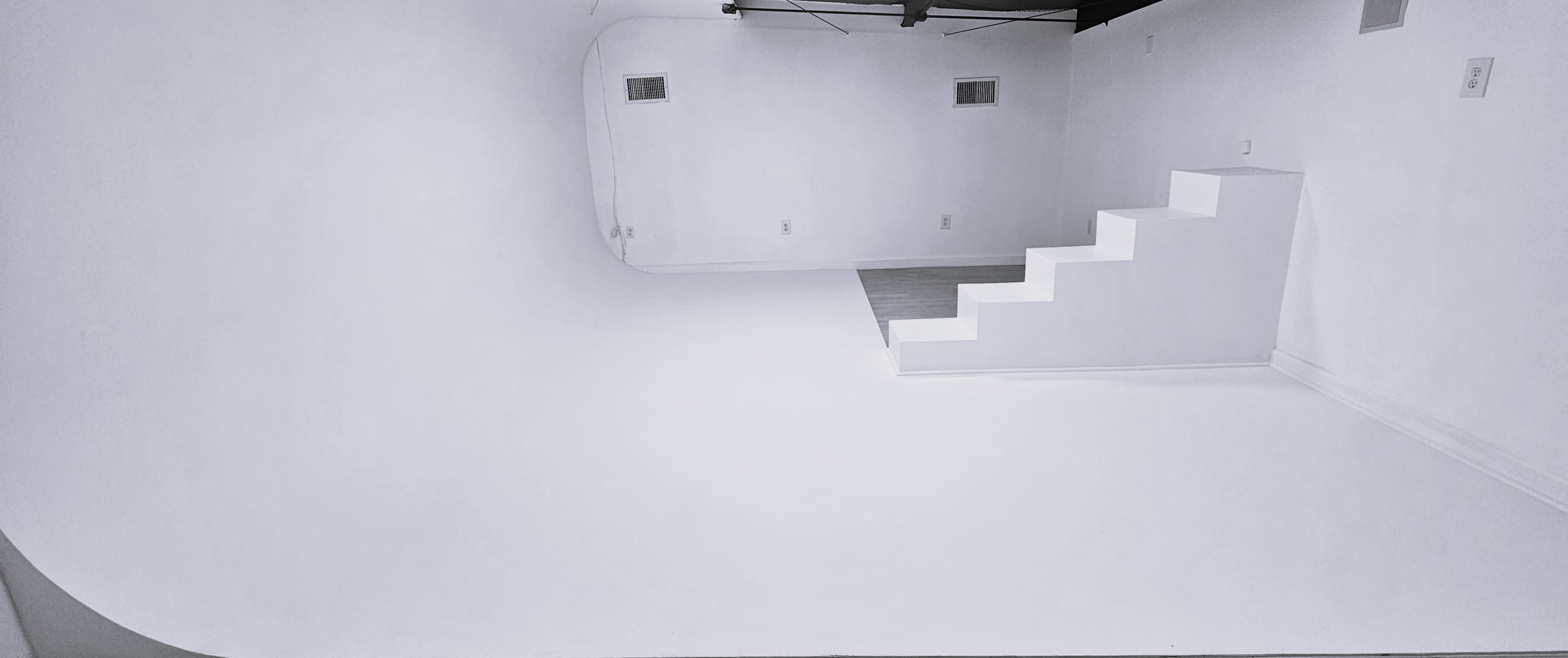 The image shows a minimalist white room featuring a curved wall, a staircase with three steps, and a section of a gray floor.