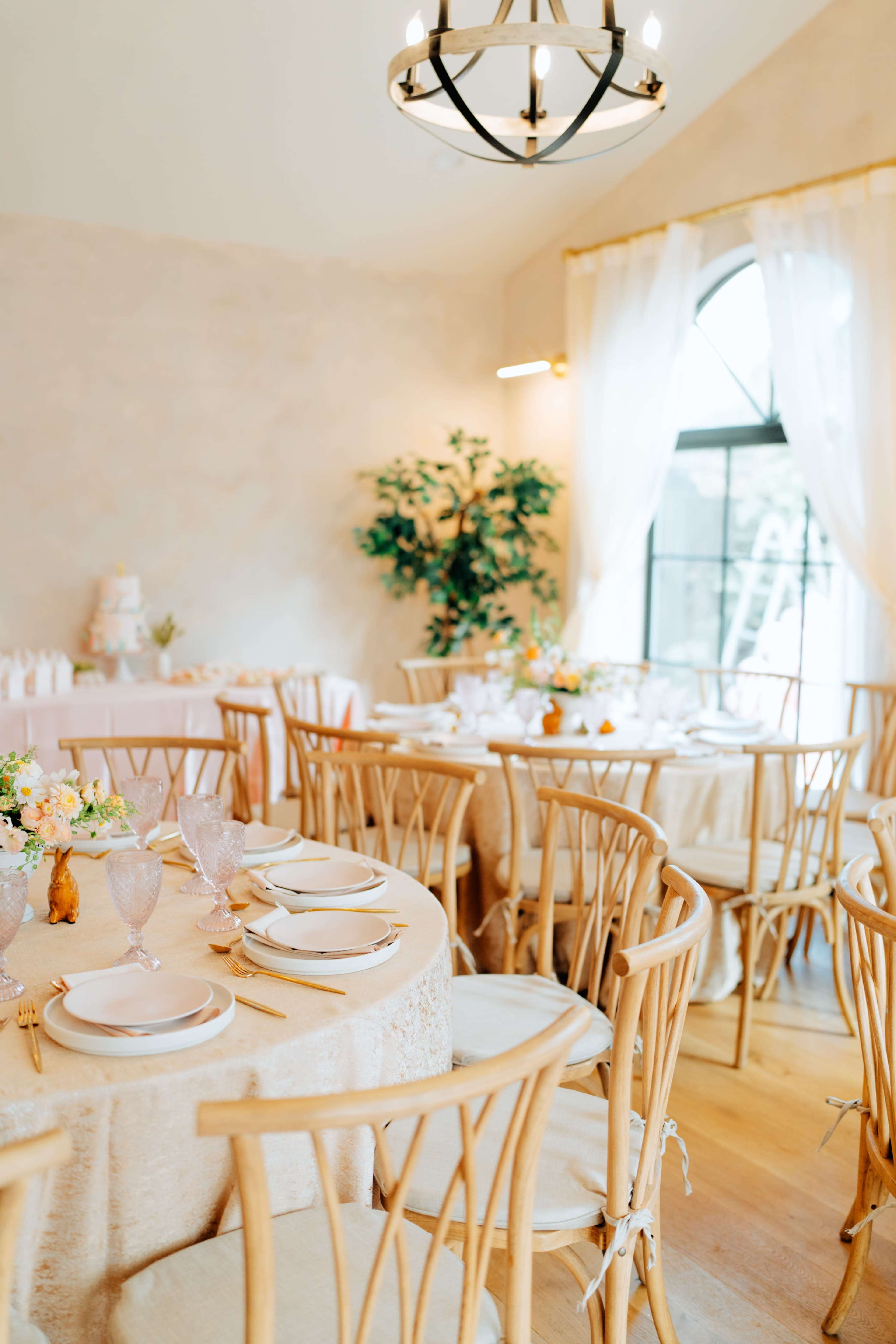 The image shows a elegantly decorated dining area with wooden chairs, round tables covered in light fabric, and floral centerpieces.