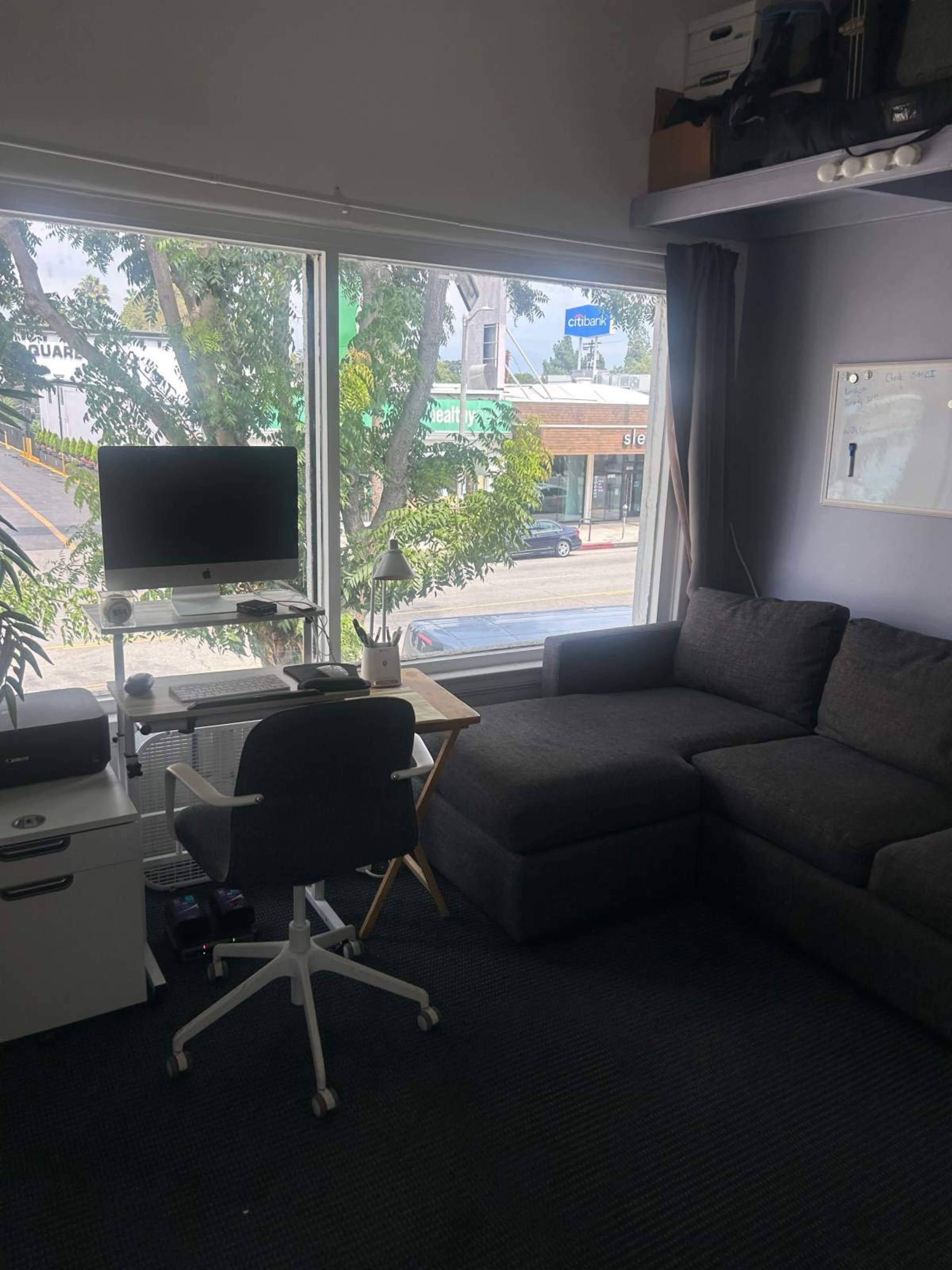 The image shows a small living area with a grey sectional sofa, a desk with a computer, and a window overlooking a street lined with trees and storefronts.