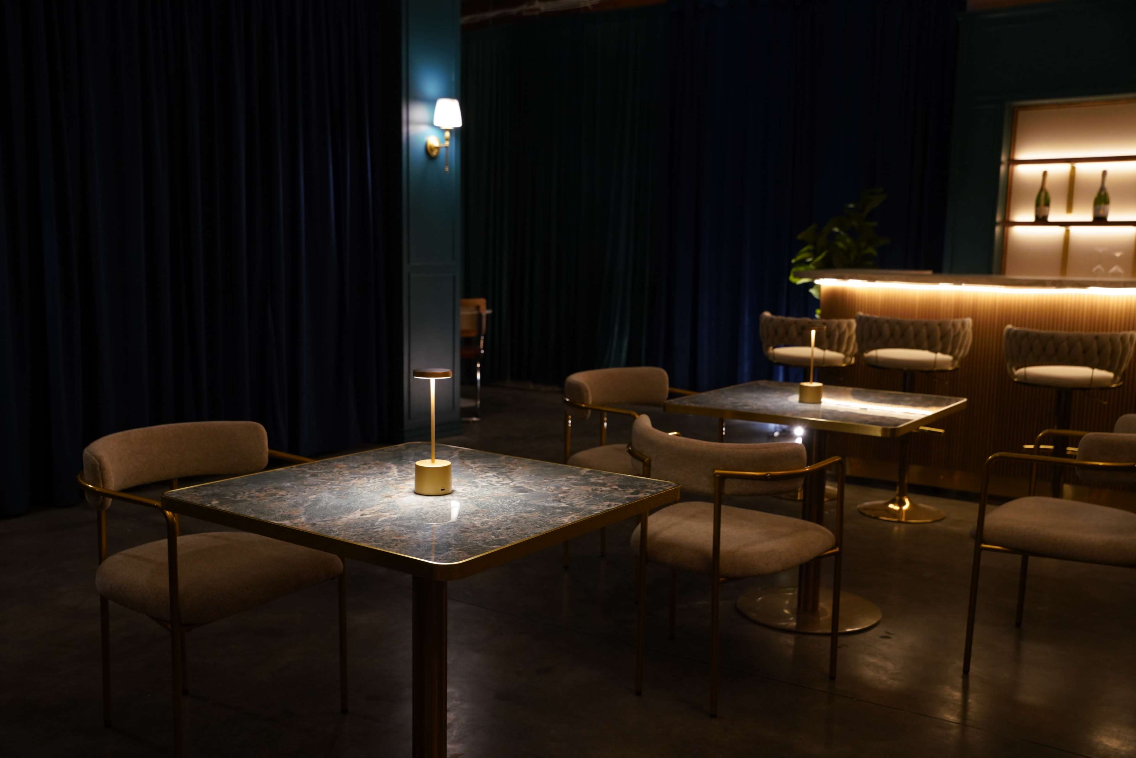 The image shows a dimly lit lounge area with several tables featuring marble tops and gold accents, surrounded by dark blue curtains and a softly illuminated bar in the background.