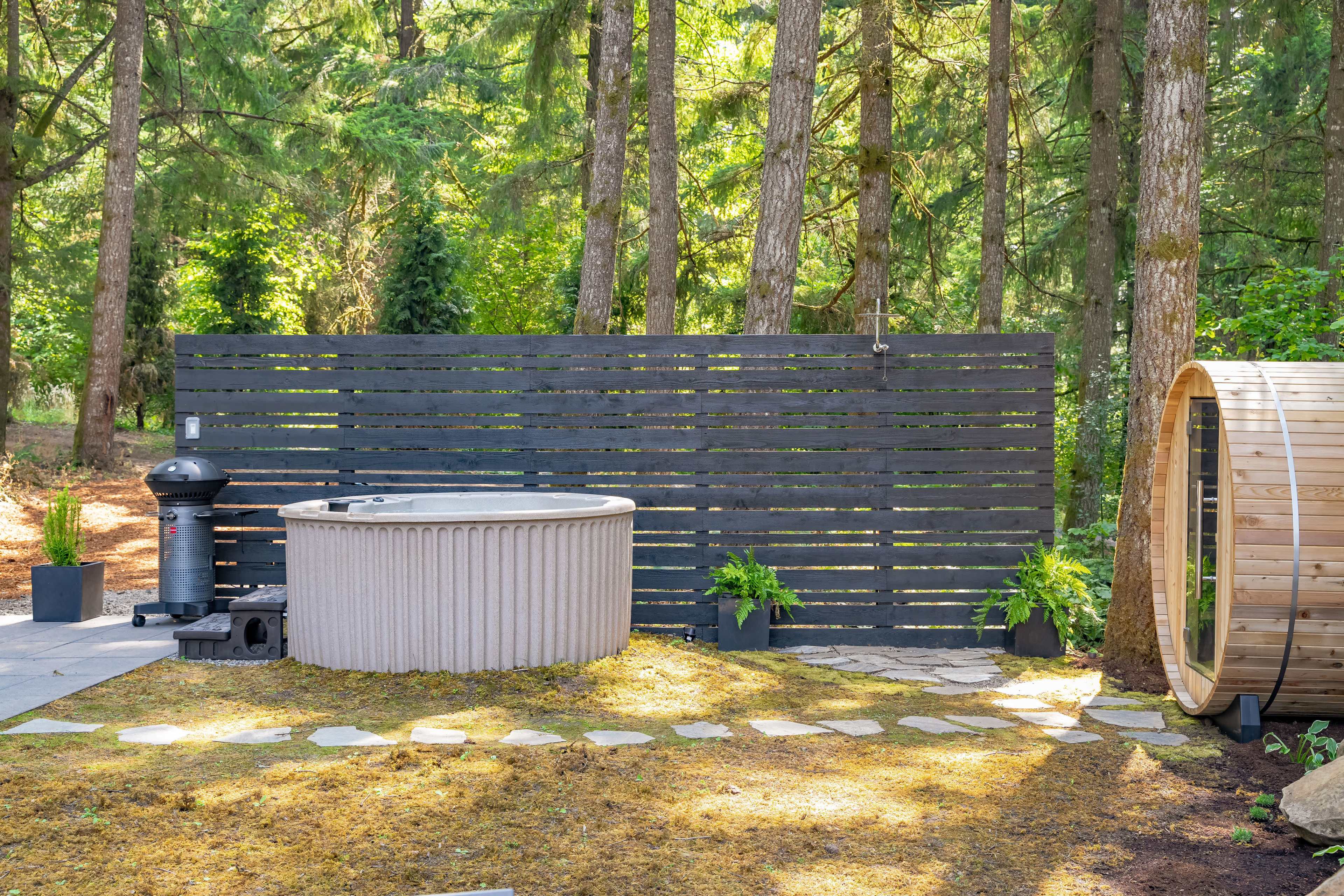A landscaped outdoor area featuring a hot tub, a barrel sauna, and a wooden privacy screen, surrounded by trees and stone pathways.