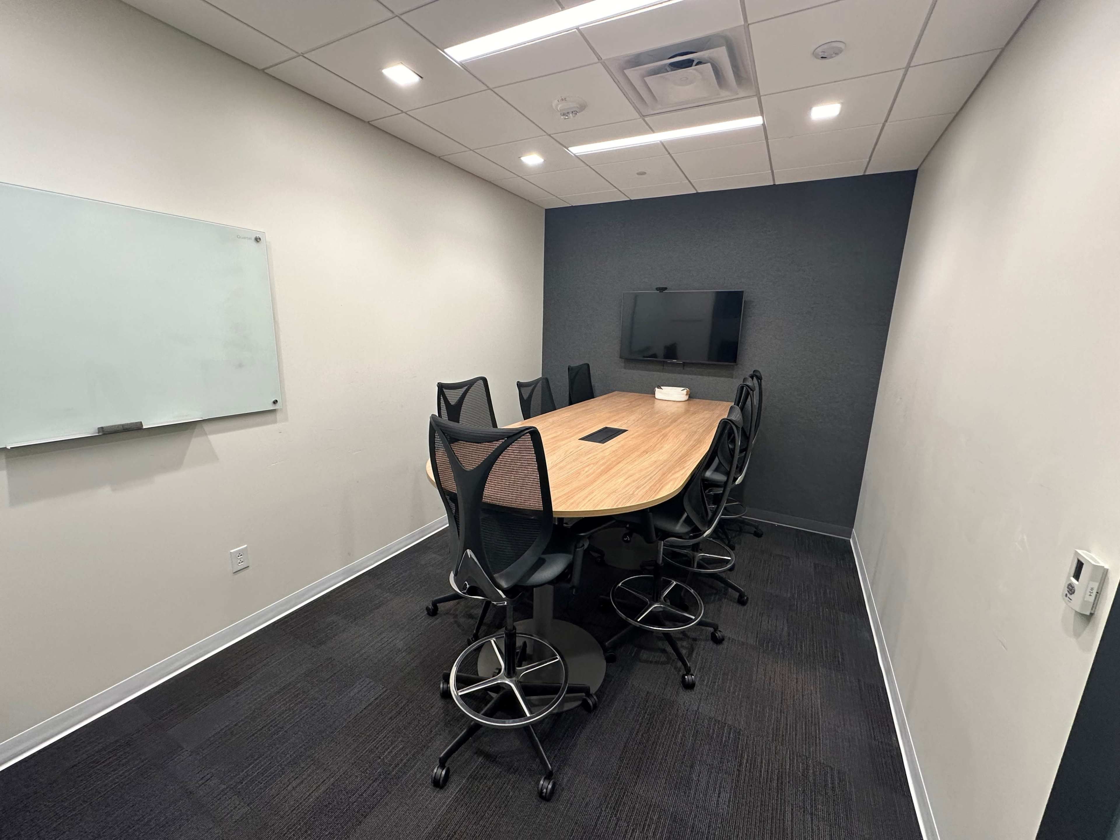 A meeting room features a large wooden table surrounded by six black ergonomic chairs, a wall-mounted TV, and a whiteboard.