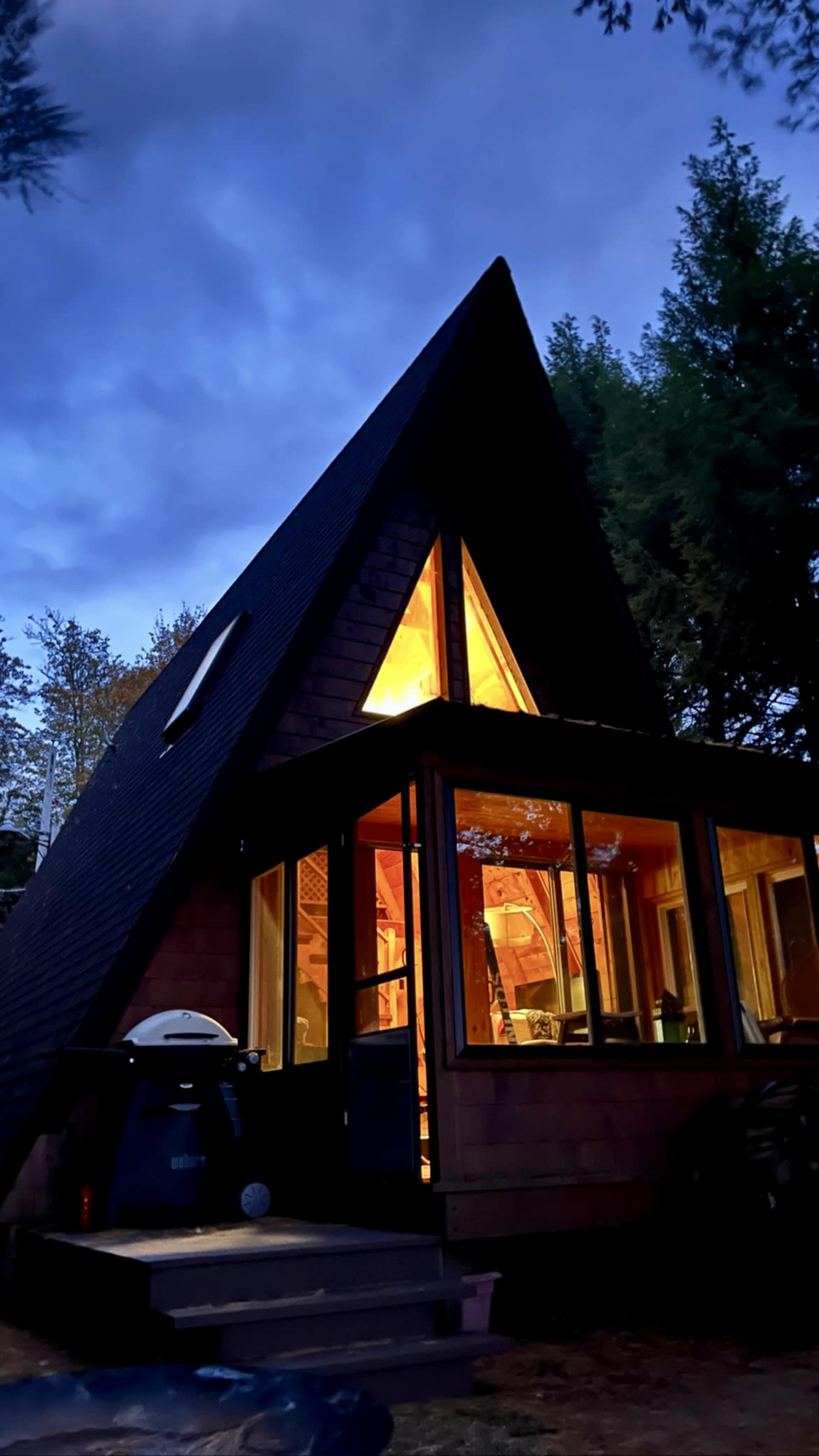 A triangular-roofed cabin with large windows is illuminated from within, surrounded by trees under a twilight sky.