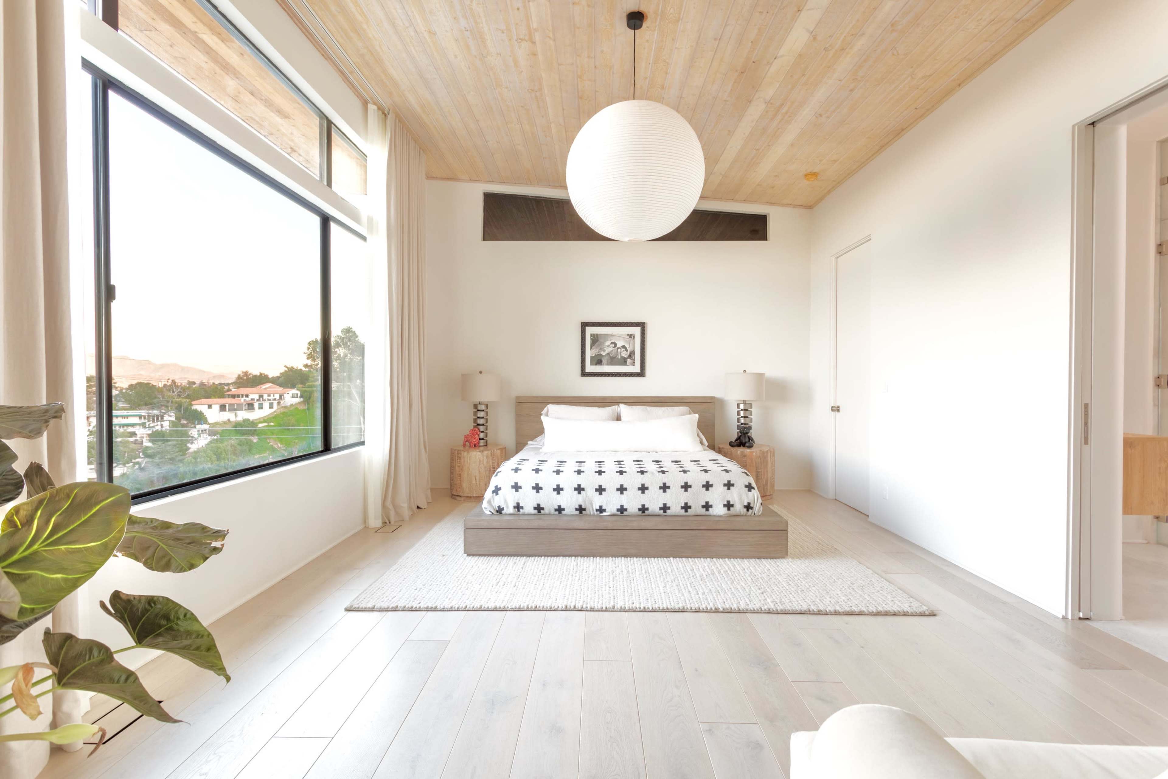 A spacious bedroom features a large bed with a patterned duvet, minimalist furniture, and a large window offering a view of the outdoors.