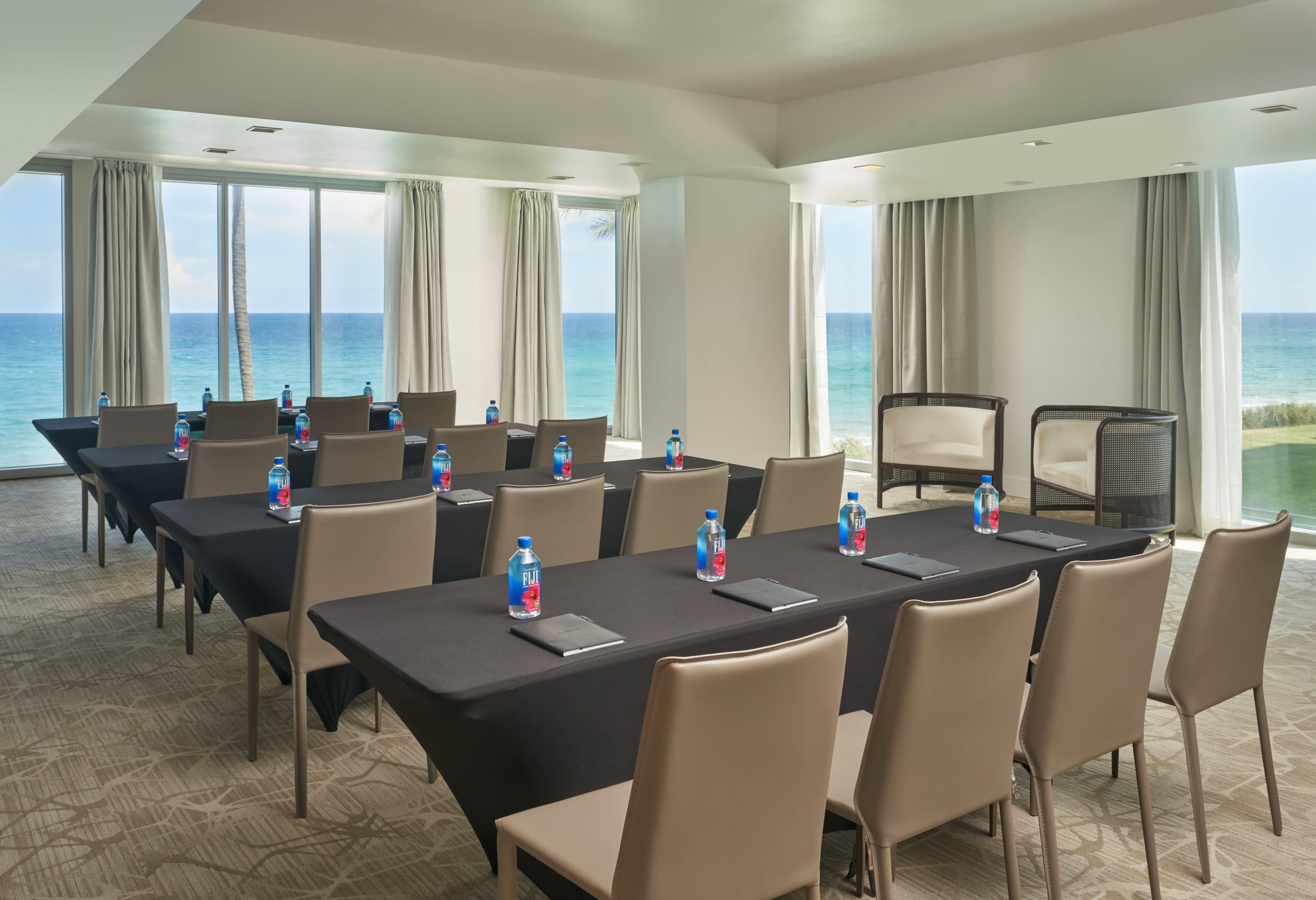 A meeting room is arranged with facing tables and chairs, featuring water bottles and notepads, with large windows showcasing an ocean view.