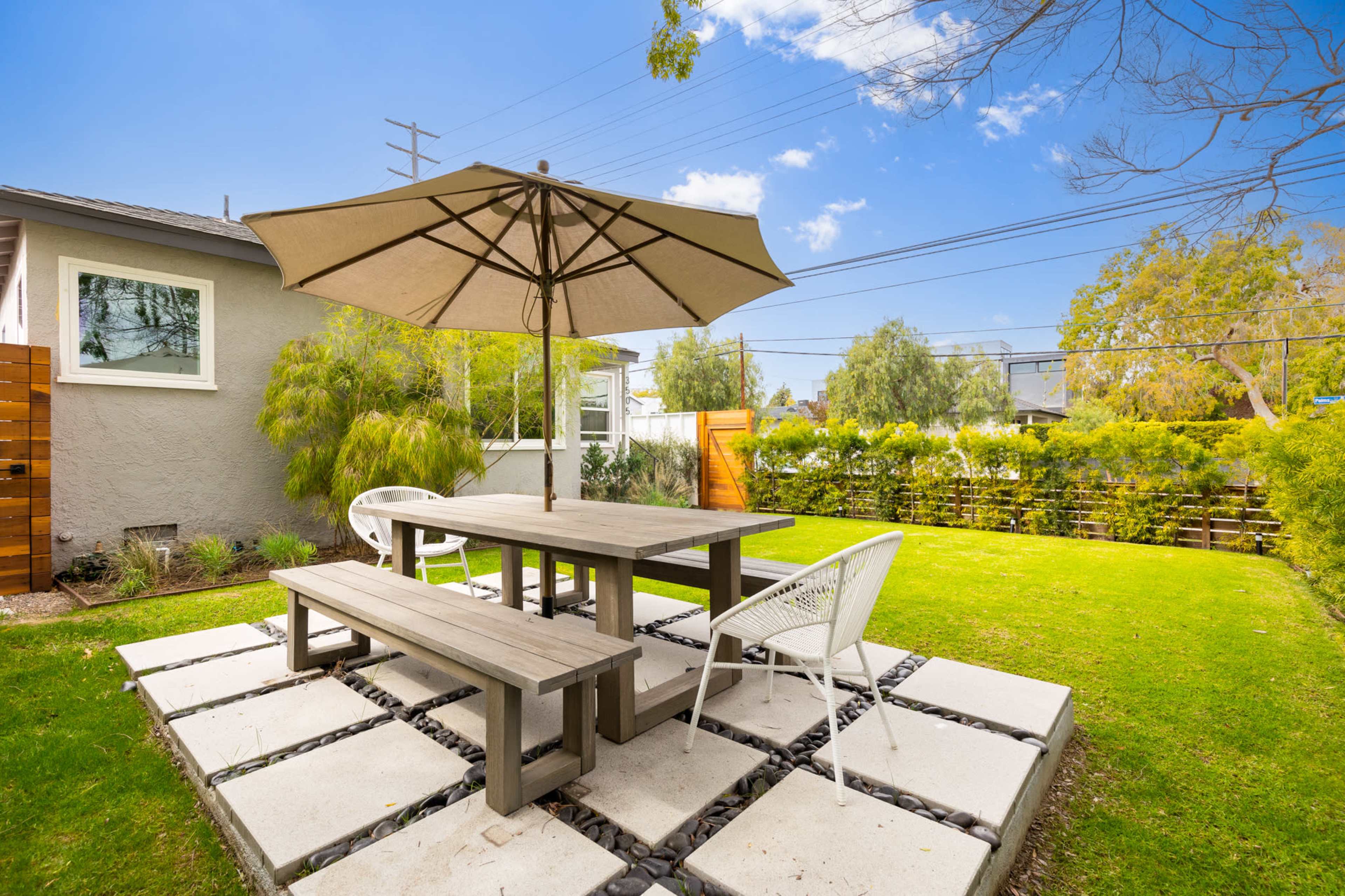 A patio area features a table with benches and a white umbrella, surrounded by green grass and decorative landscaping.