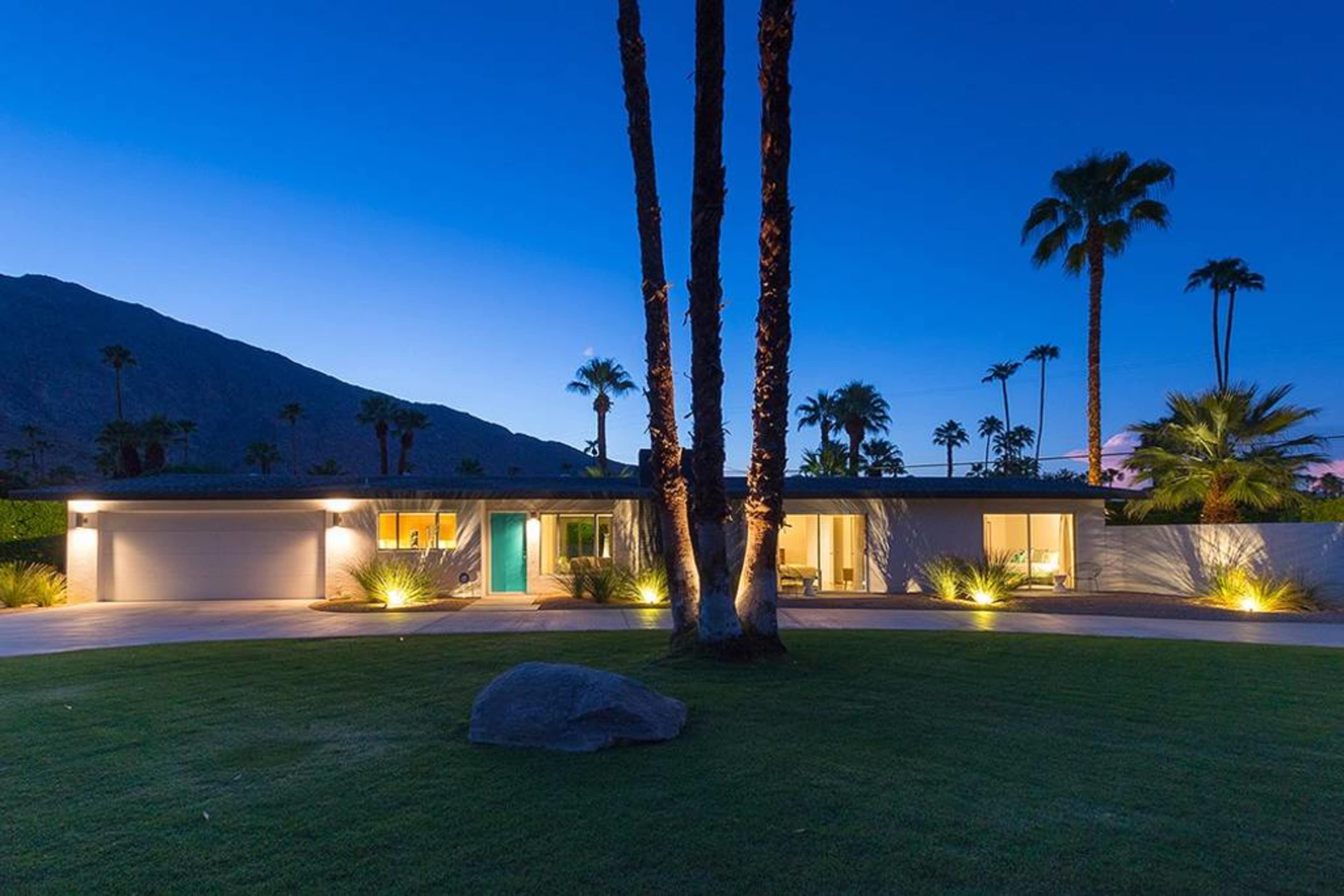 A modern single-story house is illuminated at dusk, surrounded by palm trees and set against a mountain backdrop.