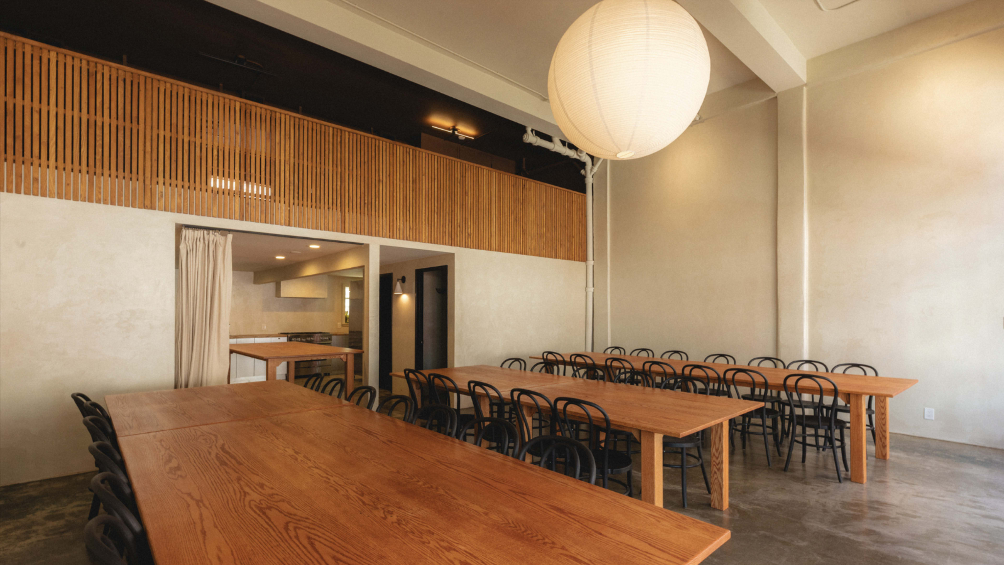 The image shows a spacious dining area with long wooden tables and black chairs, illuminated by a large round pendant light.