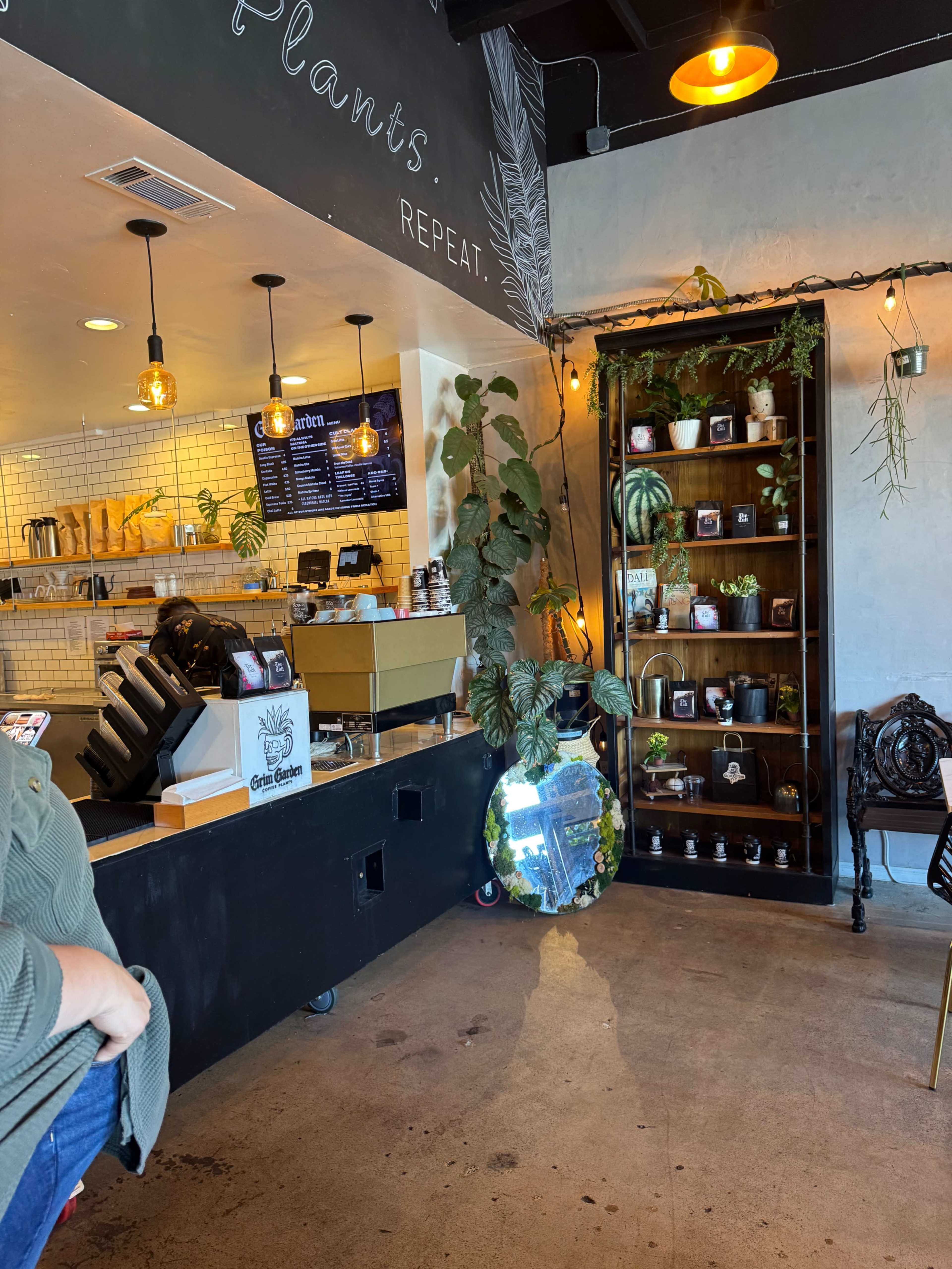 The image shows the interior of a café with a wooden counter, menu board, plants on display, and a bookshelf filled with various items.