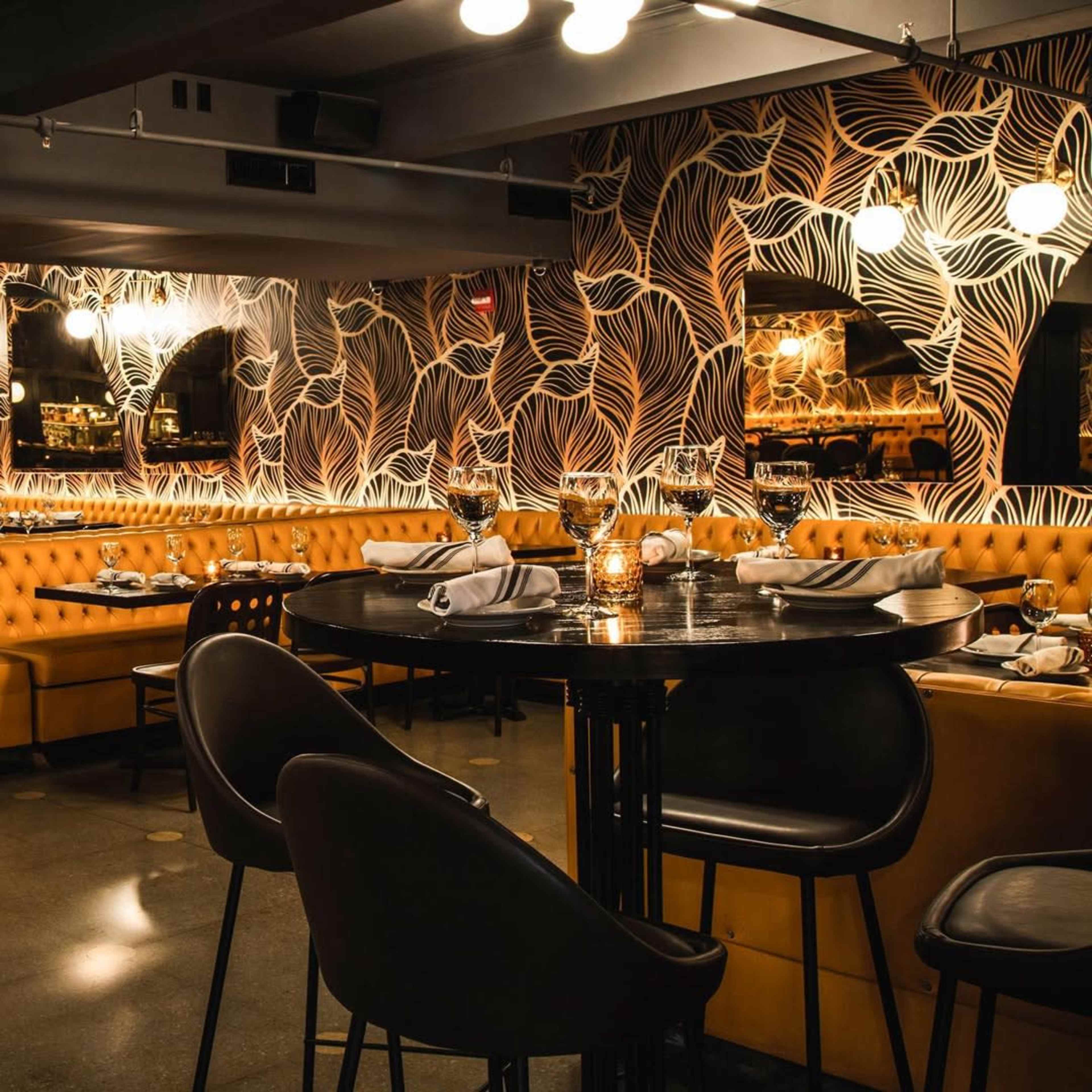 The image shows a stylish restaurant interior featuring yellow upholstered seating, patterned wallpaper, and elegant table settings with dim lighting.