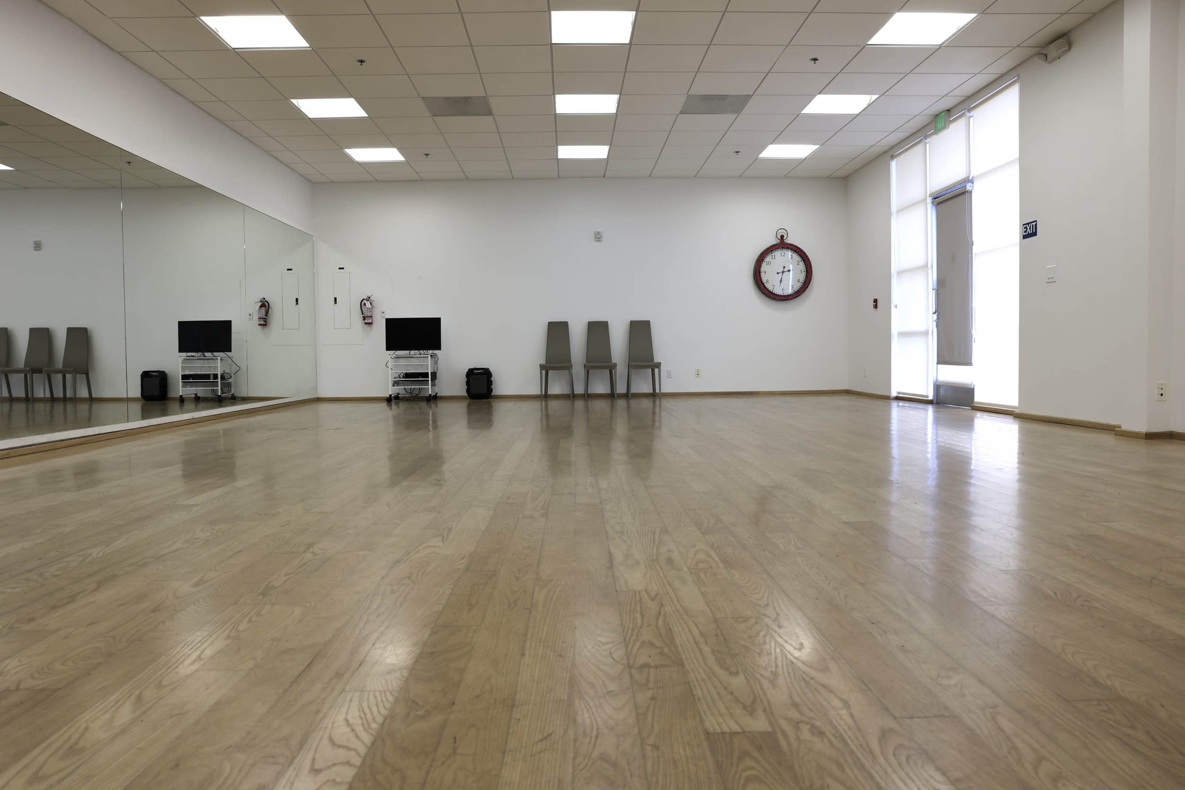 A spacious, empty room with a wooden floor, two chairs, two screens, and a wall clock, featuring a large mirror on one side.