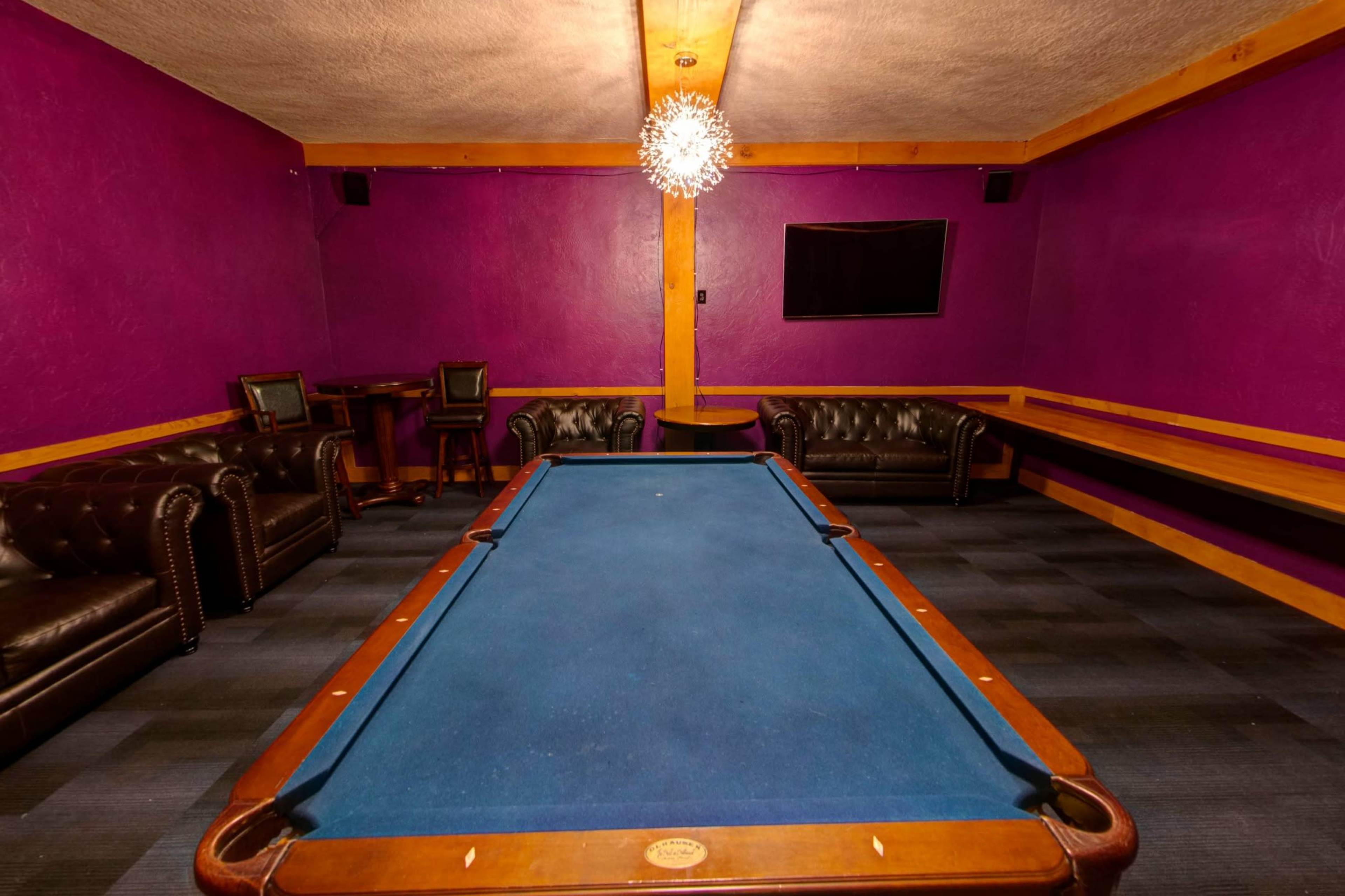 The image shows a game room featuring a blue billiard table, dark leather couches, and purple walls with a chandelier overhead.