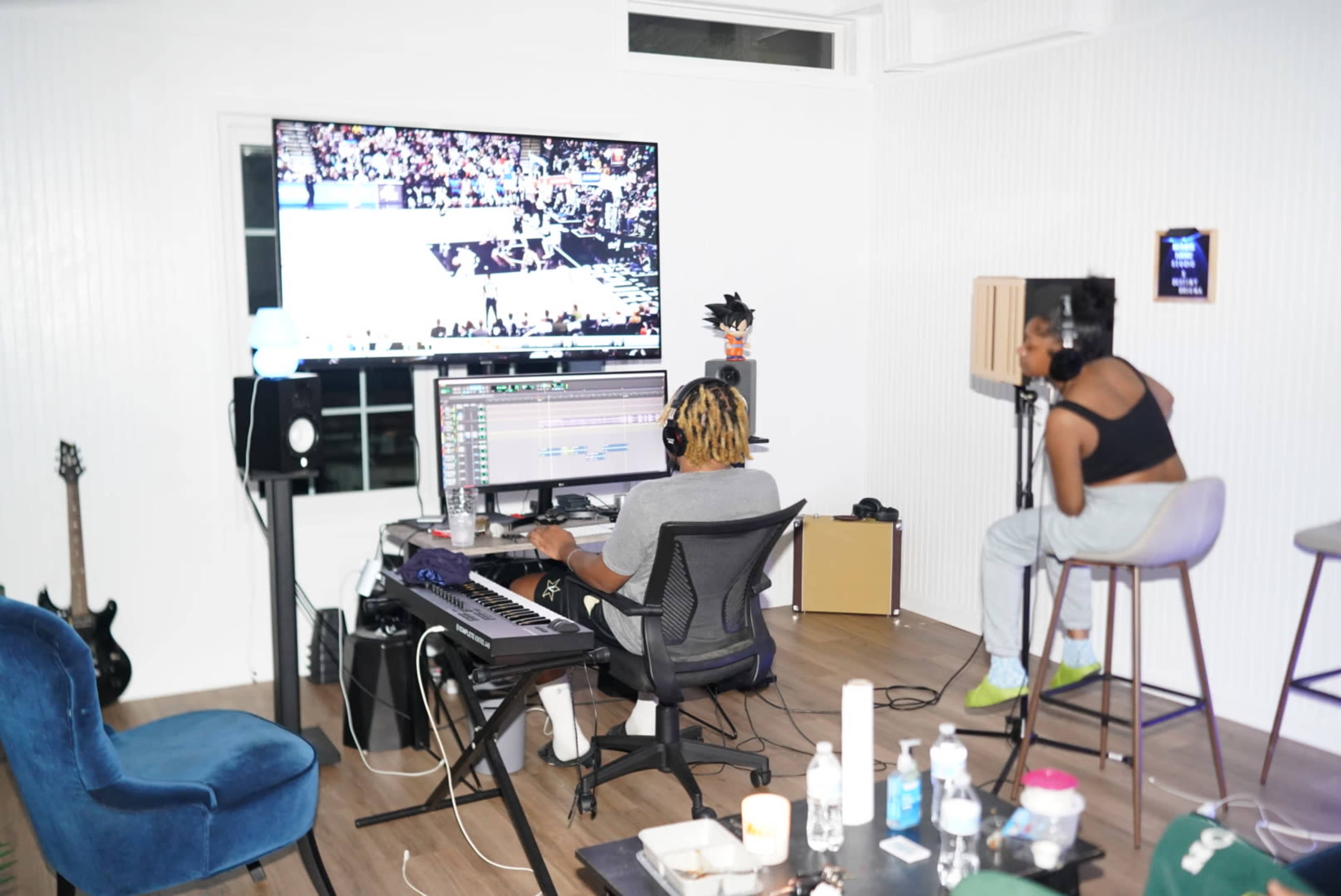 A person is working at a laptop with music software in a home studio while another person sits nearby, watching a basketball game on a large screen.