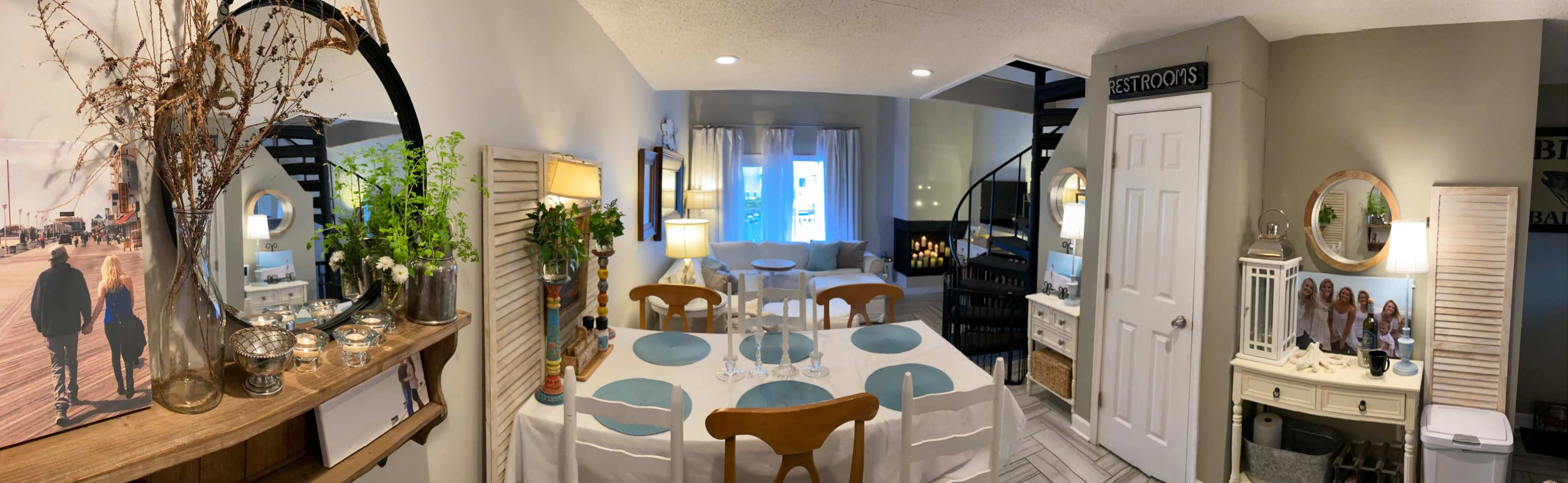 A cozy interior space with a dining area featuring a table set for a meal, a staircase leading to an upper level, and various decorative elements, including plants and artwork.