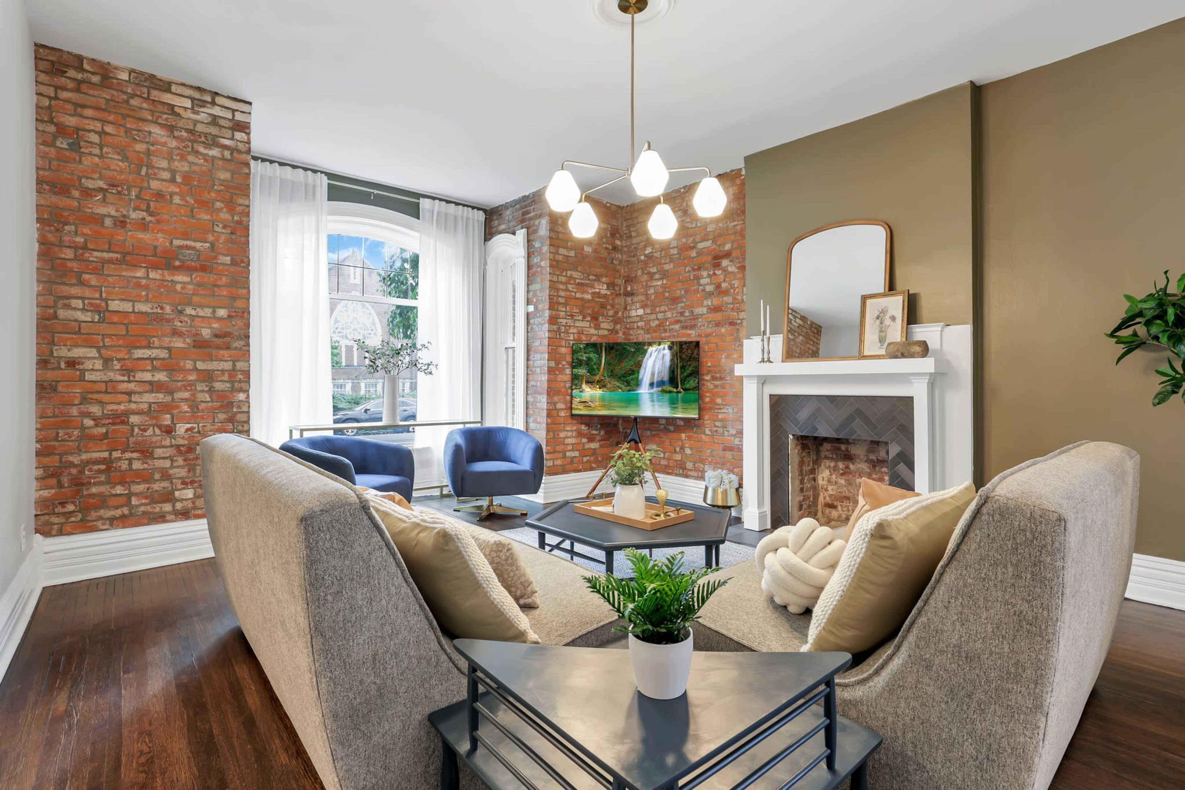 A cozy living room features a gray sofa, a coffee table, and a fireplace, with exposed brick walls and a large window displaying a view of the outdoors.