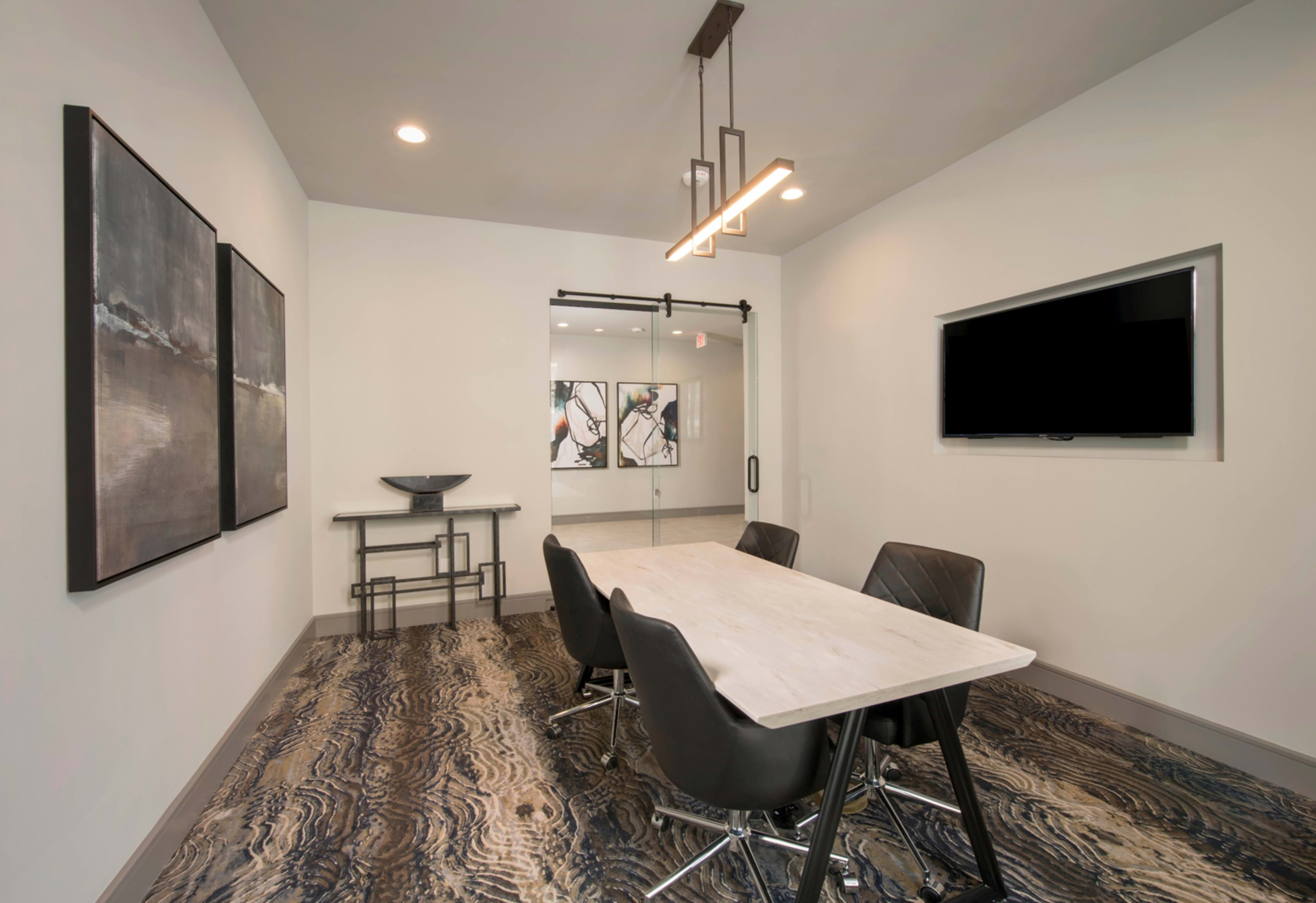 A modern conference room features a marble table surrounded by black chairs, with artwork on the walls and a television mounted opposite the entrance.