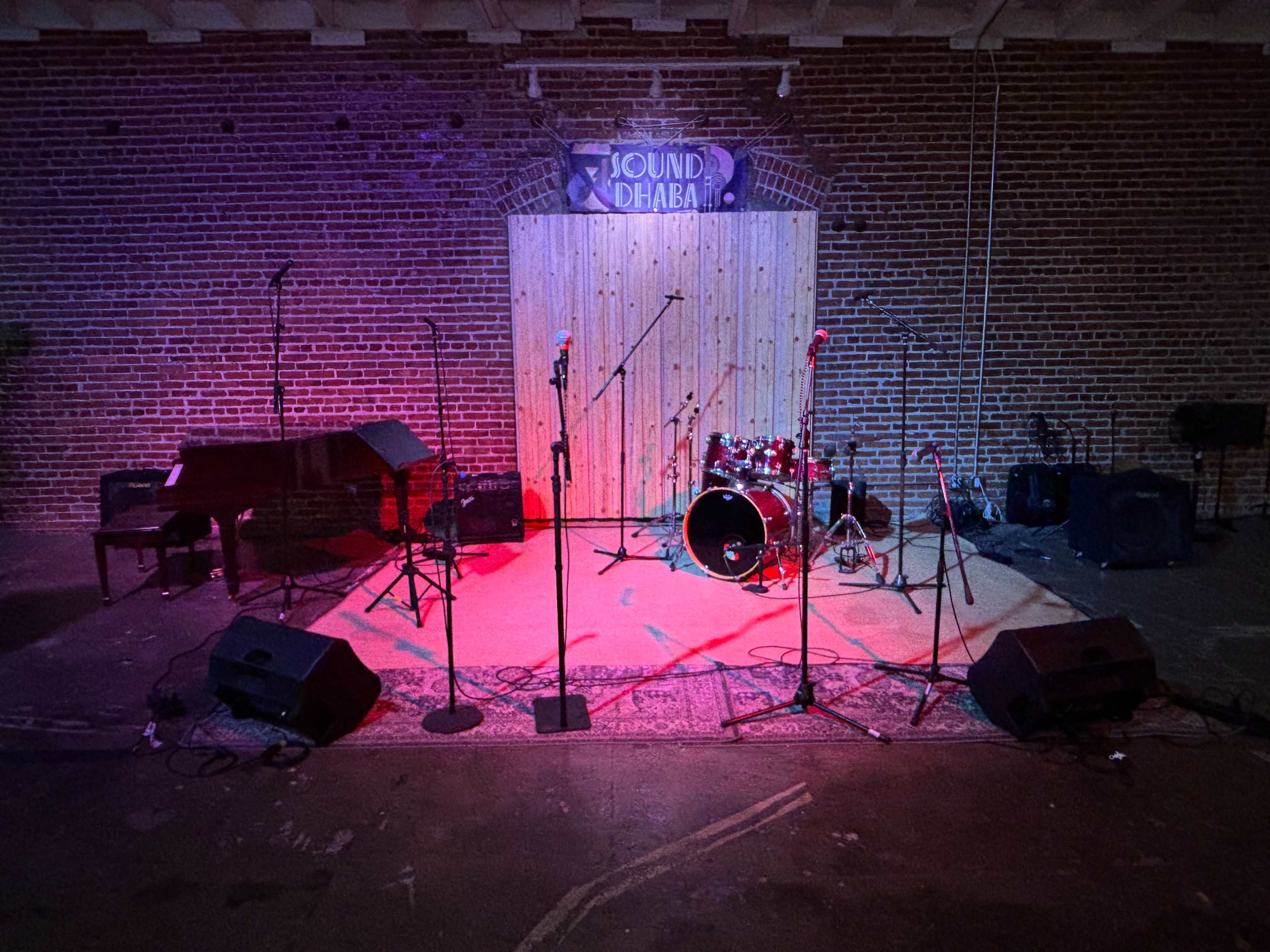 The image shows an empty stage setup with musical instruments, including a piano and a drum set, surrounded by microphone stands and speakers, against a brick wall backdrop.