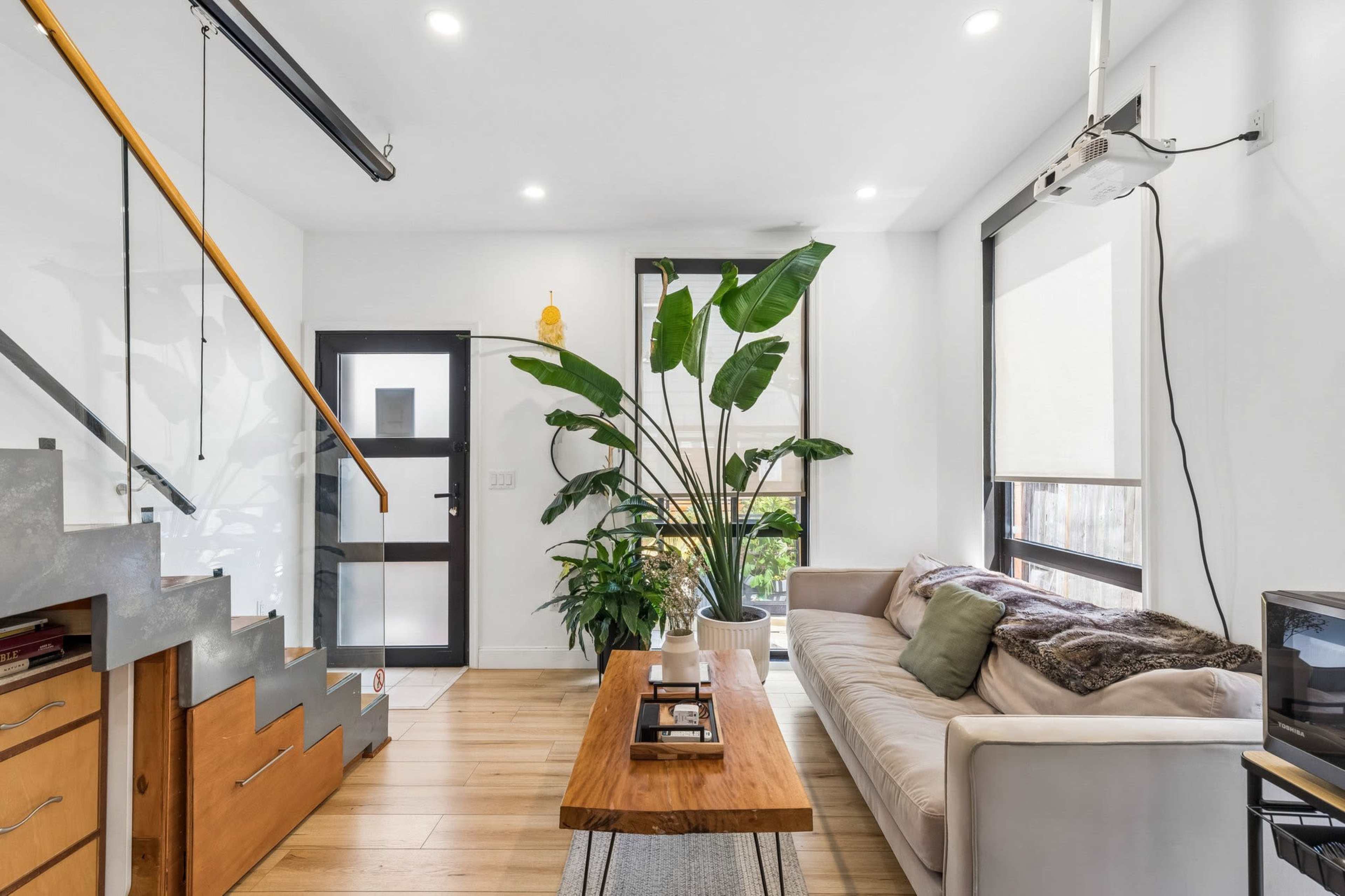 The image shows a modern living room with a staircase, a potted plant, a sofa, a wooden coffee table, and a front door.